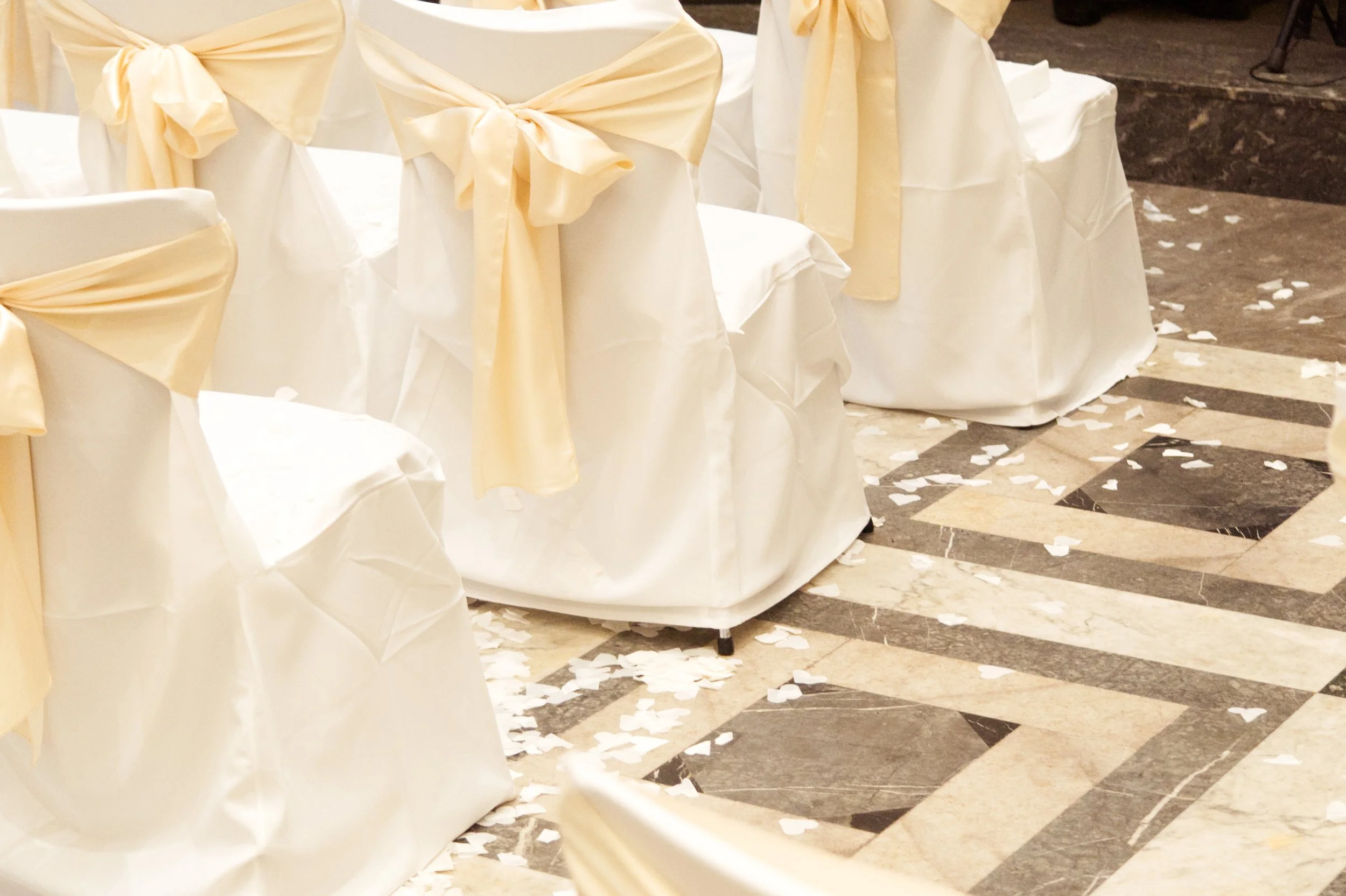 Stühle mit weißen Überzügen und cremefarbenen Schleifen, auf dem Boden verstreute weiße Konfettis, vermutlich für eine Hochzeit oder Feier vorbereitet.