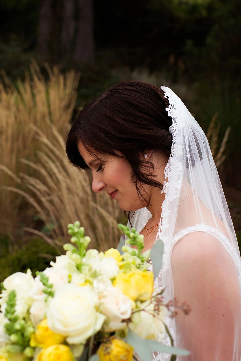 Braut mit dunklem Haar in Brautkleid und Schleier, die einen Blumenstrauß hält, im Freien bei Sonnenuntergang.