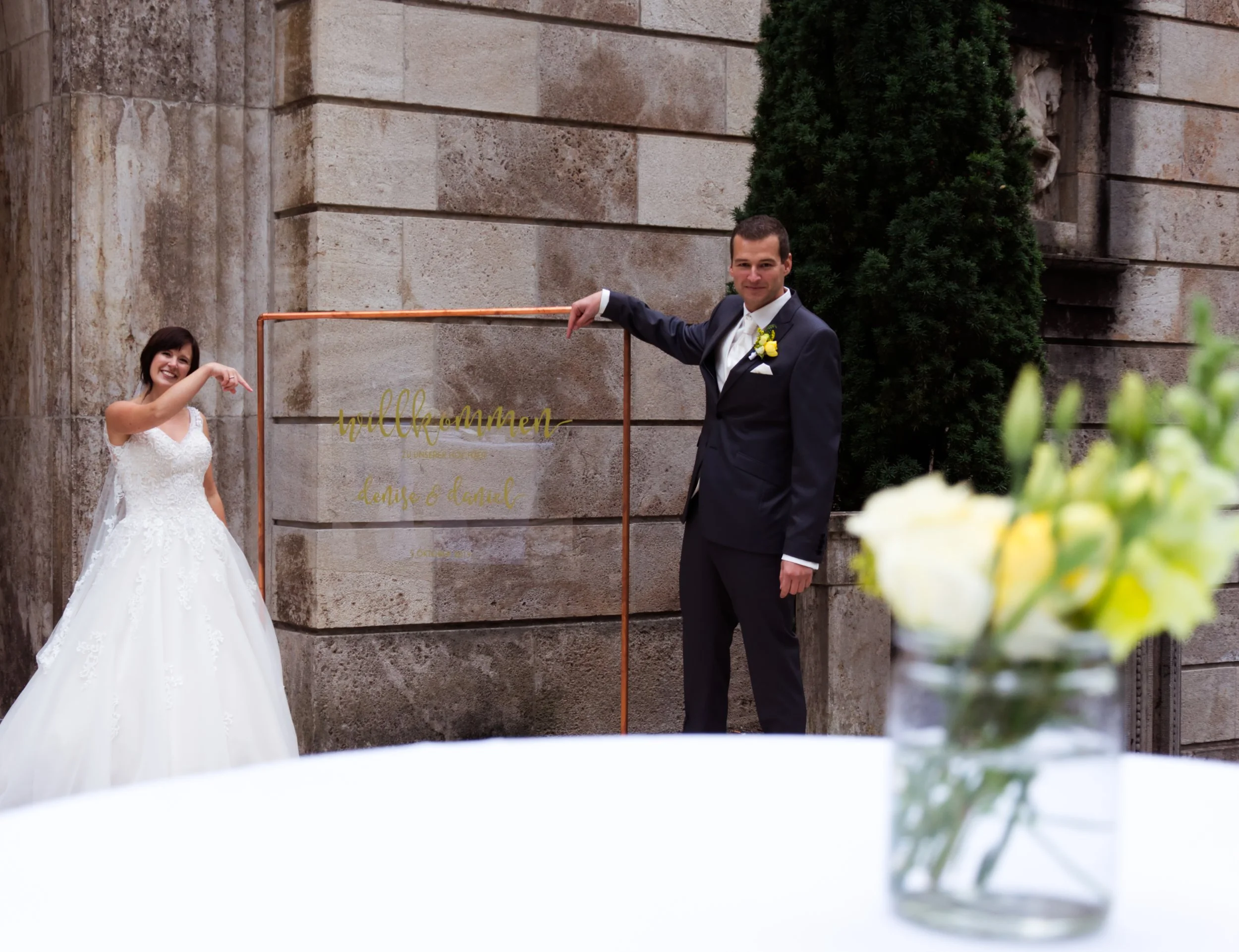 Braut und Bräutigam posieren bei einer Hochzeit, im Hintergrund eine Steinmauer, im Vordergrund unscharf eine Vase mit weißen und gelben Blumen.