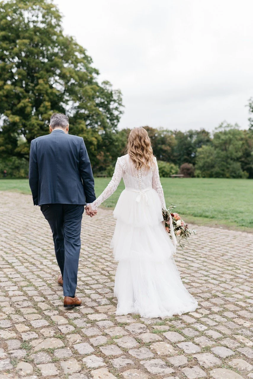 Ein Brautpaar hält Händchen beim Spaziergang auf einem gepflasterten Weg im Grünen, die Braut trägt ein weißes Hochzeitskleid und hält einen Blumenstrauß, während der Bräutigam einen dunklen Anzug trägt.