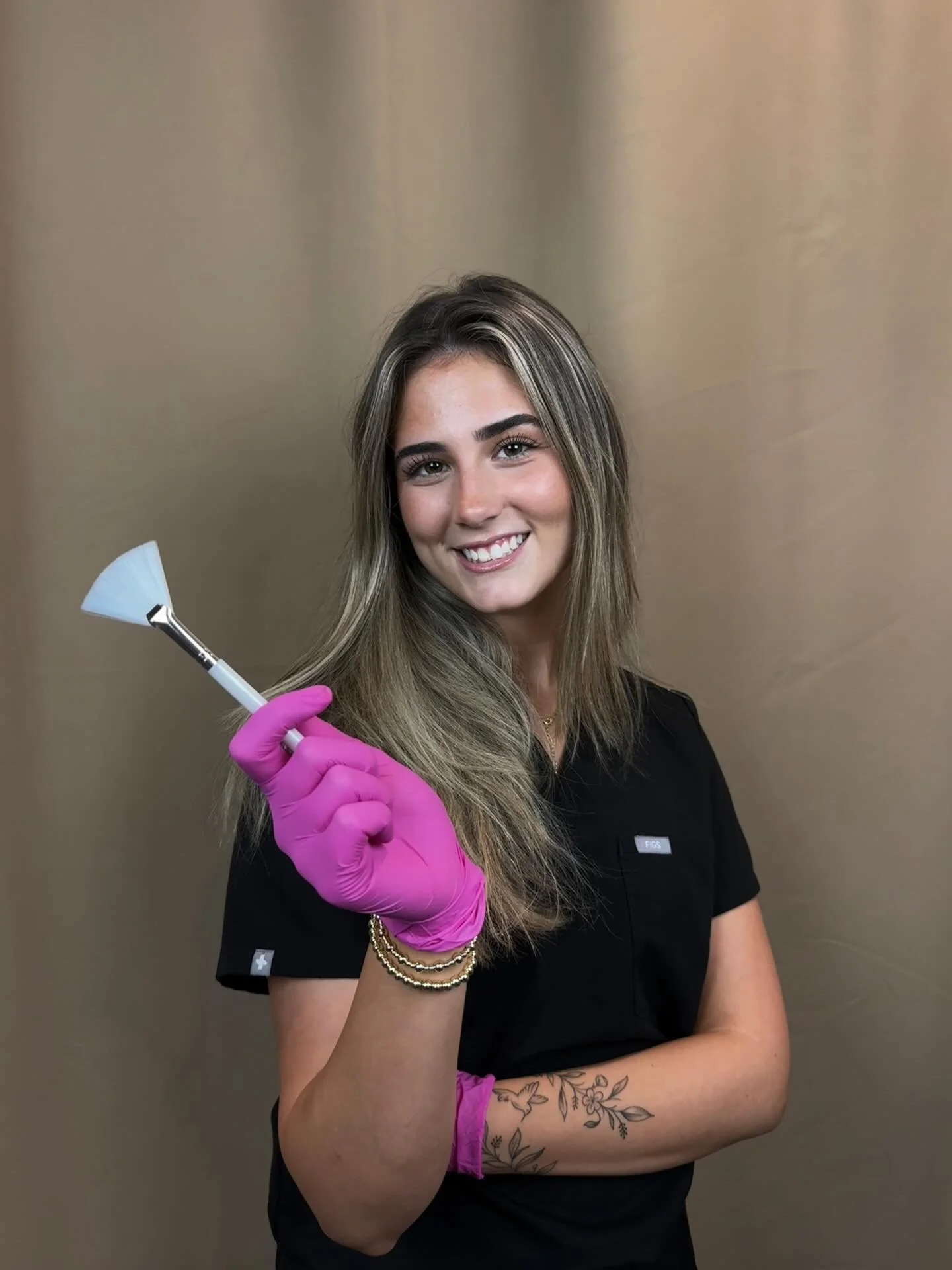 A smiling woman with long hair, wearing a black uniform and pink gloves, holding a dental tool, standing against a plain background.