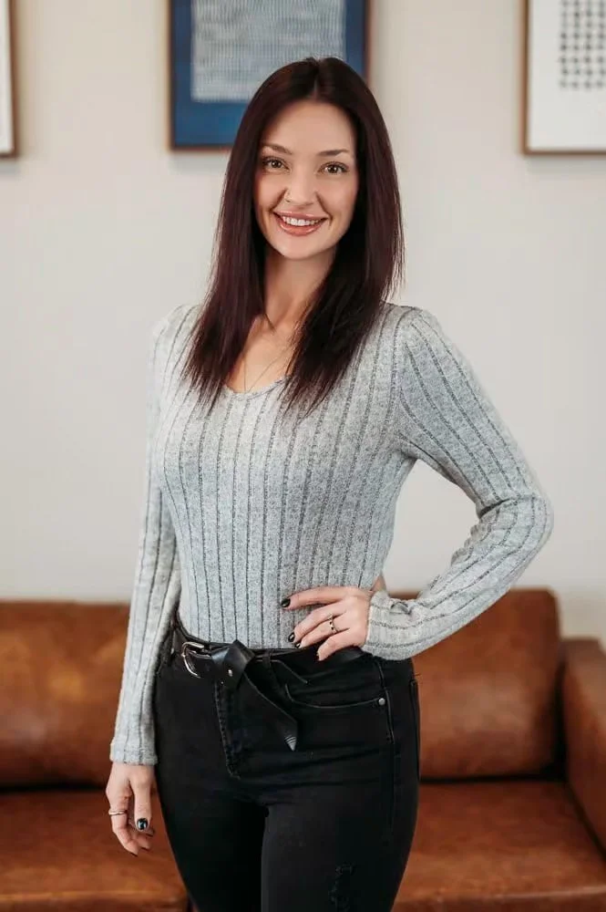 A woman with long dark hair wearing a light gray striped sweater and black pants, smiling and standing in a room with framed art on the wall behind her.