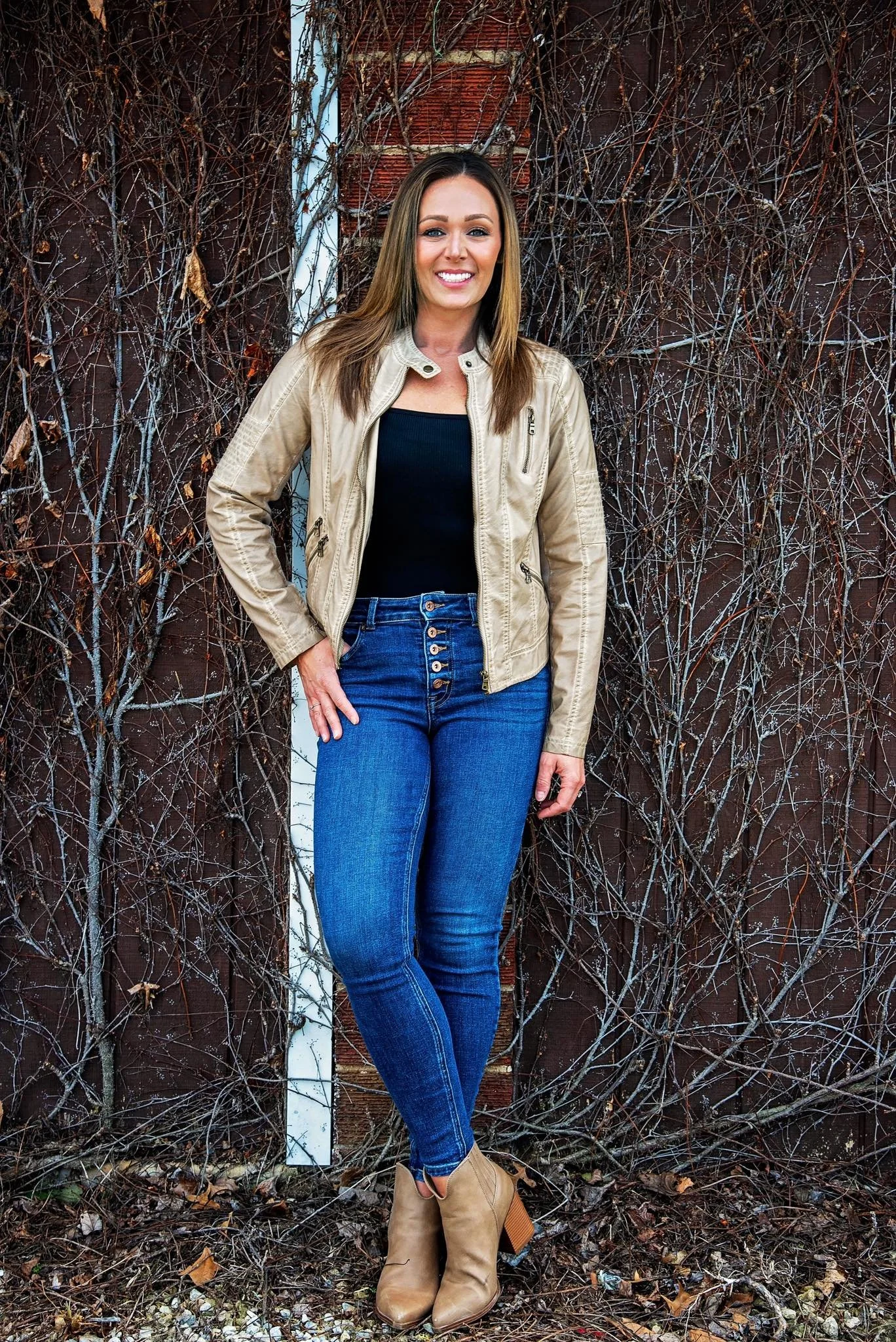 A woman with brown hair, wearing a tan leather jacket, black top, blue jeans, and tan ankle boots, standing outdoors against a wall covered with dried vines and peeling paint.