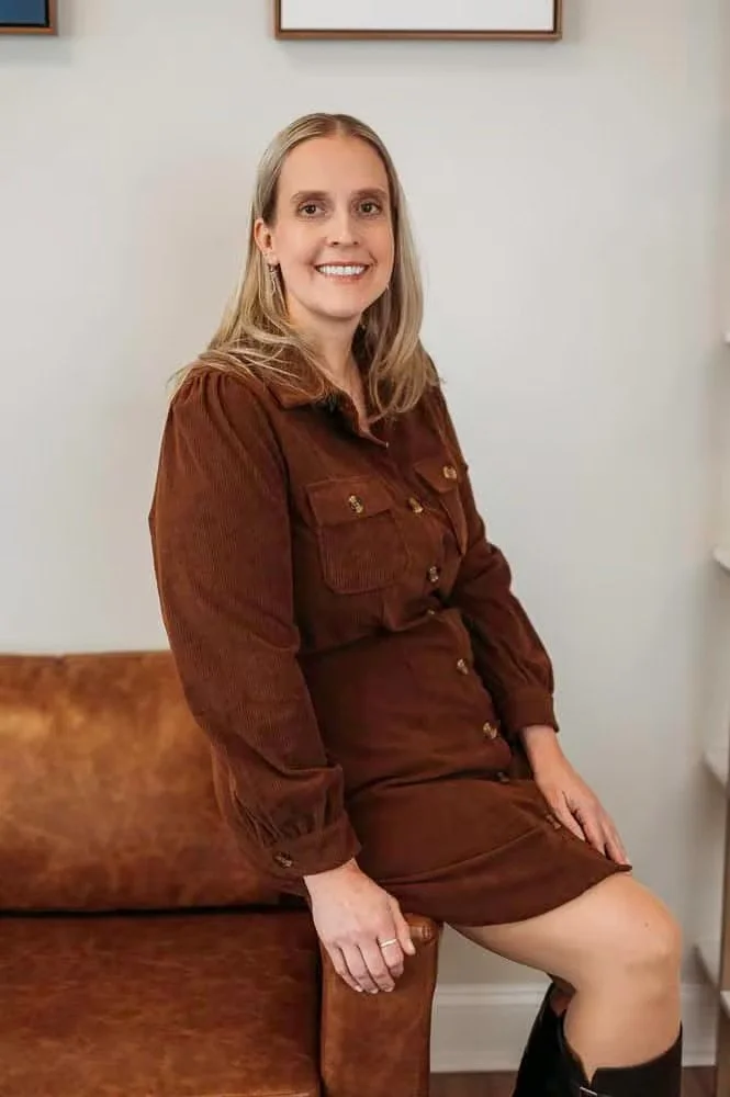 A woman with shoulder-length blonde hair and a big smile, sitting on a brown leather couch in a room with light-colored walls and a framed picture above her.
