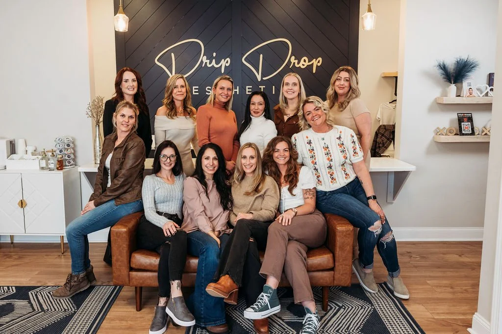 A group of fifteen women posing for a photo inside a medspa. They are sitting and standing in front of a black wall with the words 'Drip Drop Aesthetic' on it. The medspahas white and beige decor with shelves, decorations, and a small table.