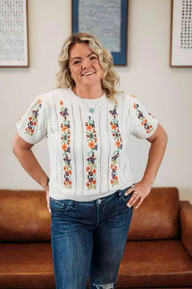 A woman with blonde, curly hair smiling, standing in a room with framed artwork on the wall behind her. She wears a white embroidered blouse and blue jeans, with one hand on her hip.
