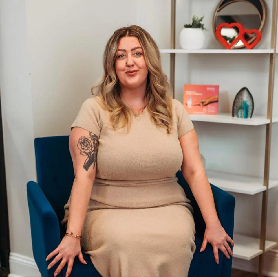 A woman with blonde hair sitting on a blue chair in a room with white shelves decorated with picture frames and ornaments.
