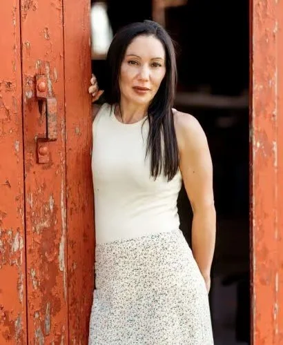 A woman with long dark hair wearing a white sleeveless top and a patterned skirt, standing between red rusty metal doors outdoors.