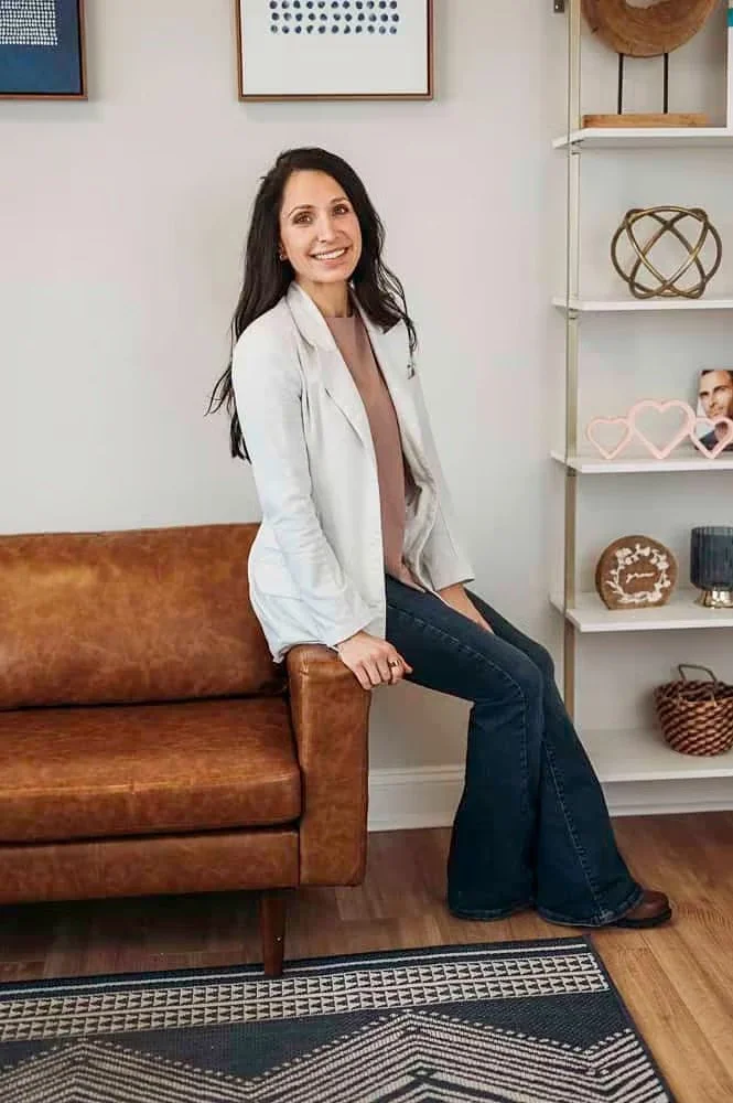 A woman with long dark hair smiling while sitting on the arm of a brown leather sofa in a modern living room with framed artwork and decorative objects on shelves.