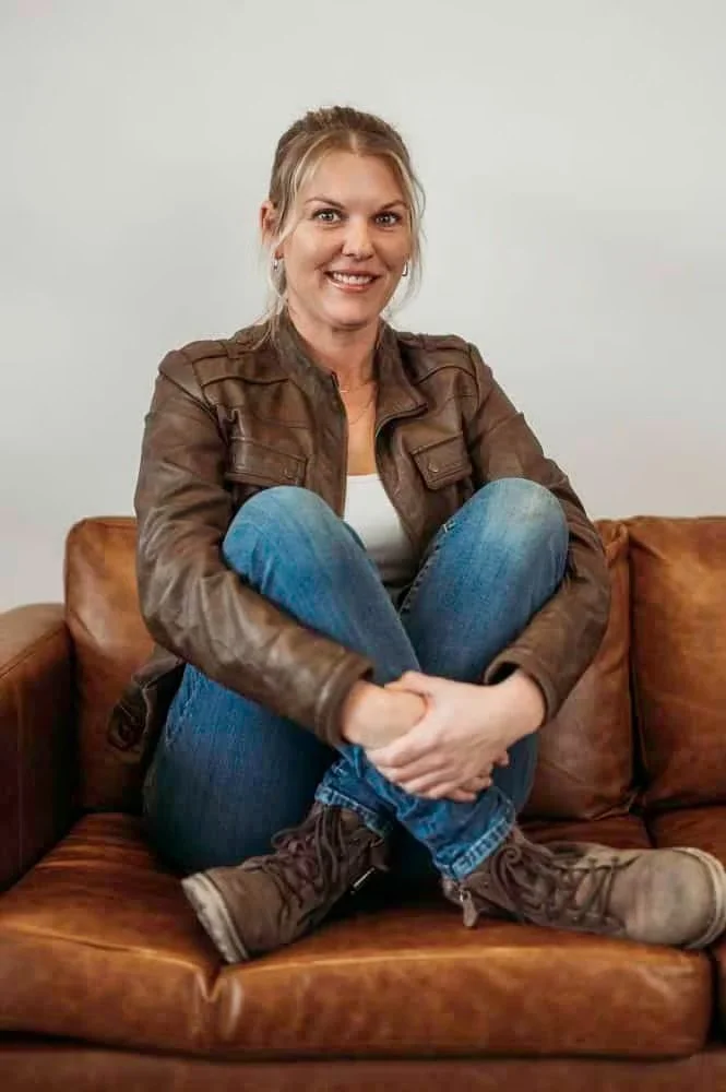 Smiling woman sitting on a brown leather sofa with her knees drawn up to her chest, wearing a brown leather jacket, white top, blue jeans, and brown boots, against a plain white wall.