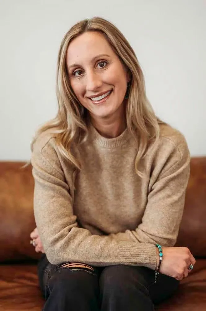 A woman with blonde hair sitting on a brown leather couch, smiling, wearing a beige sweater, and black jeans with a rip at the knee, with a white wall in the background.