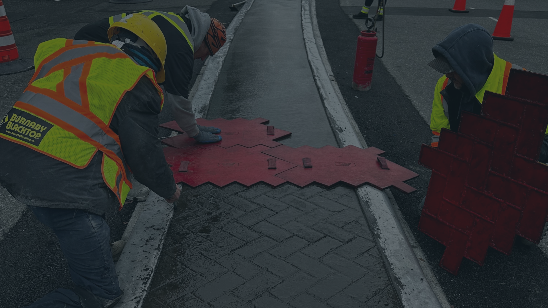 Two workers in yellow safety vests and helmets laying concrete pavers on a sidewalk, with orange traffic cones and barriers around them.