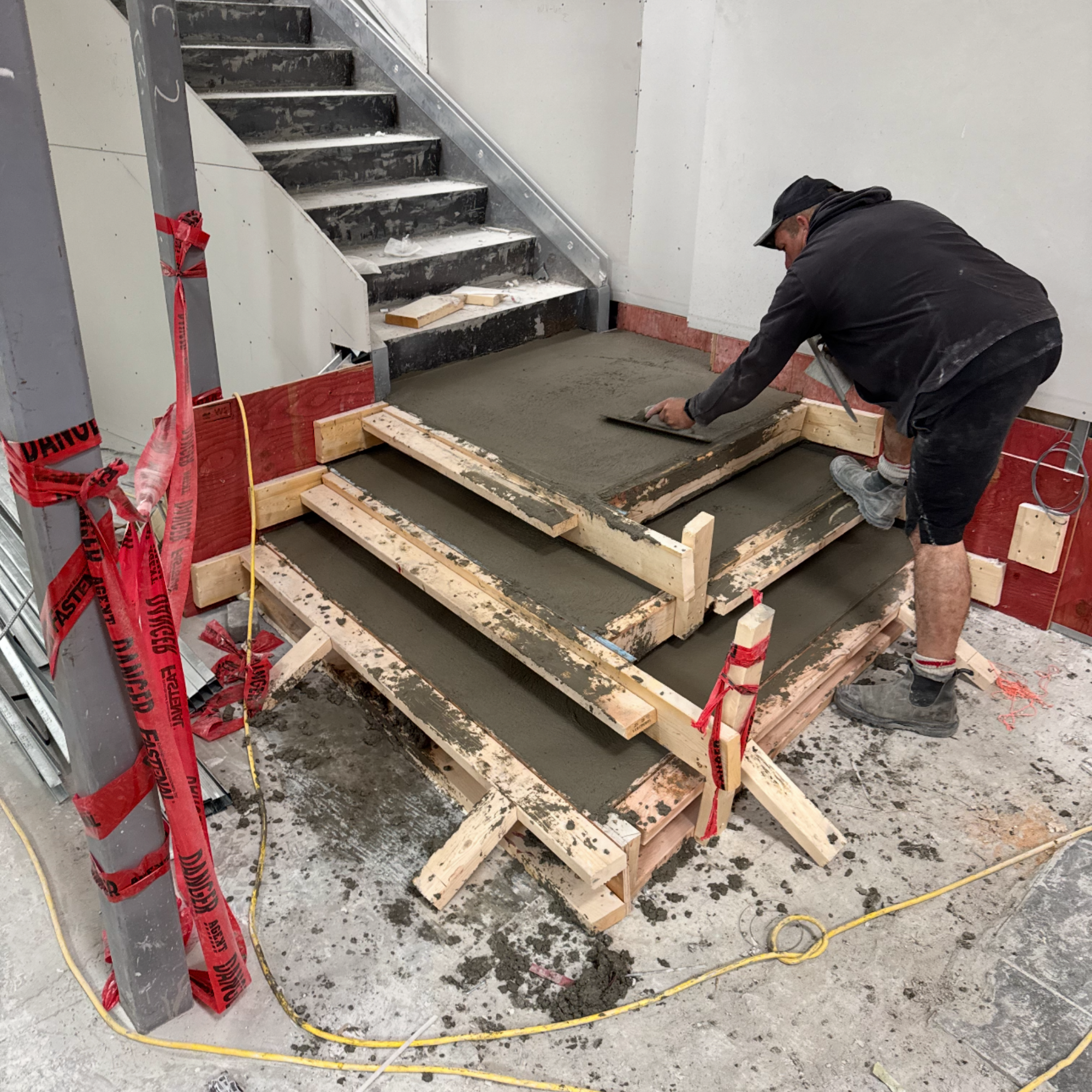 A construction worker is pouring wet concrete onto a wooden frame for a staircase landing inside a building under construction.