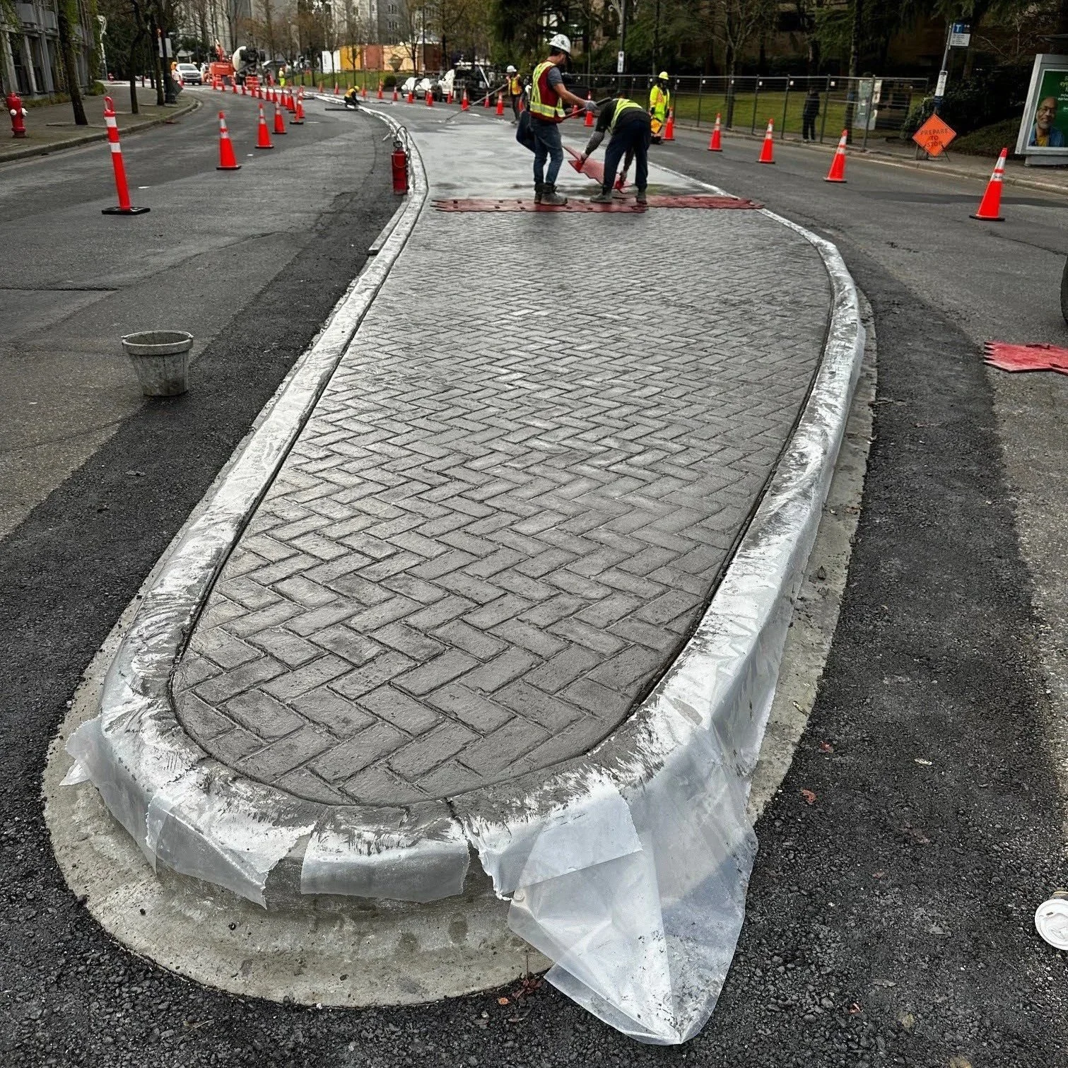 Workers laying patterned paving stones on a street, surrounded by orange traffic cones and safety barriers, with construction equipment and vehicles in the background.