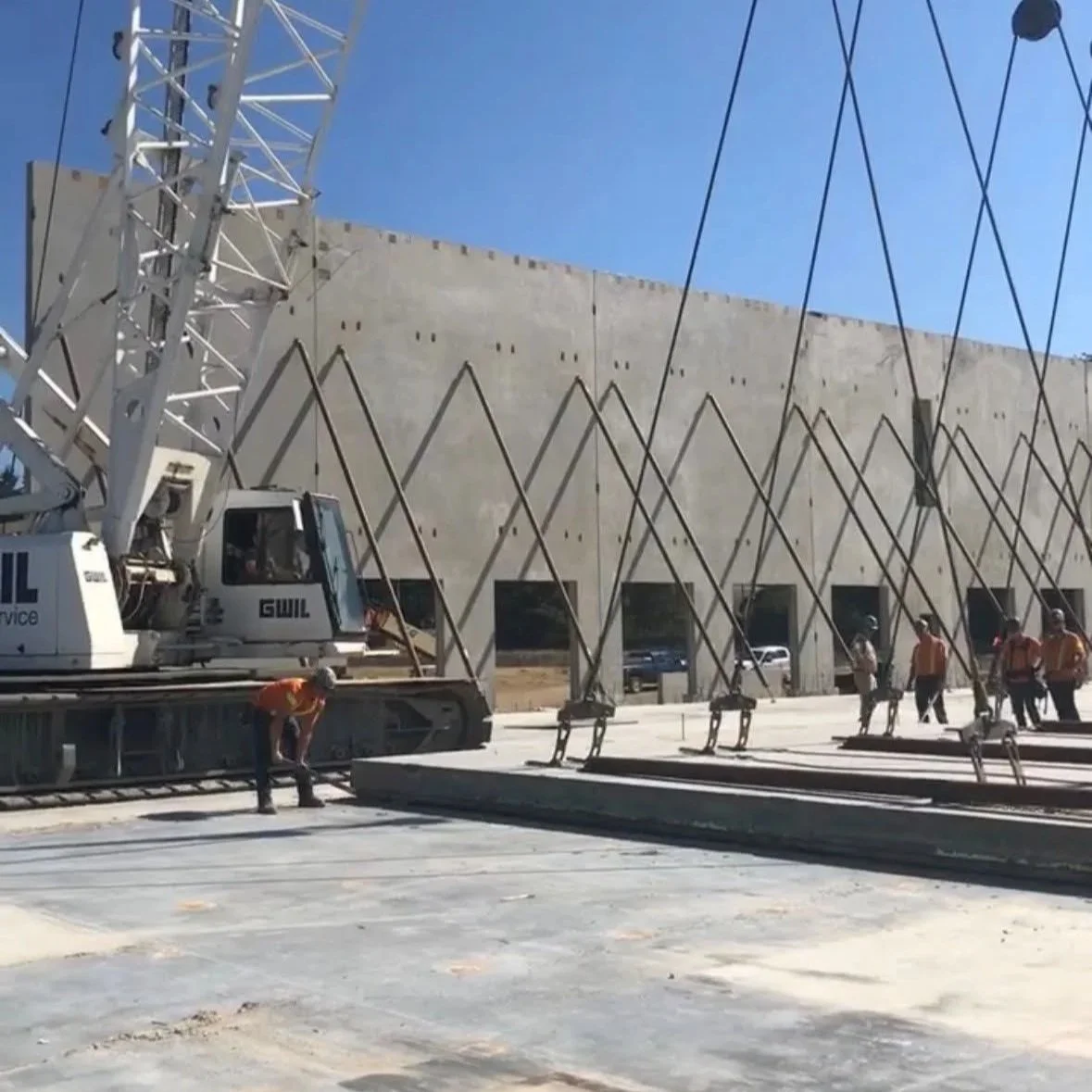Construction workers at a building site with a large crane and a concrete wall in the background. Some workers are near the ground while others walk near the building.