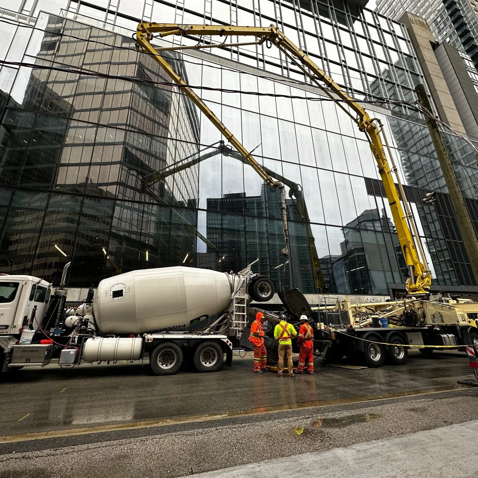 Construction workers in safety vests and helmets operate concrete pouring equipment in front of a high-rise glass building.