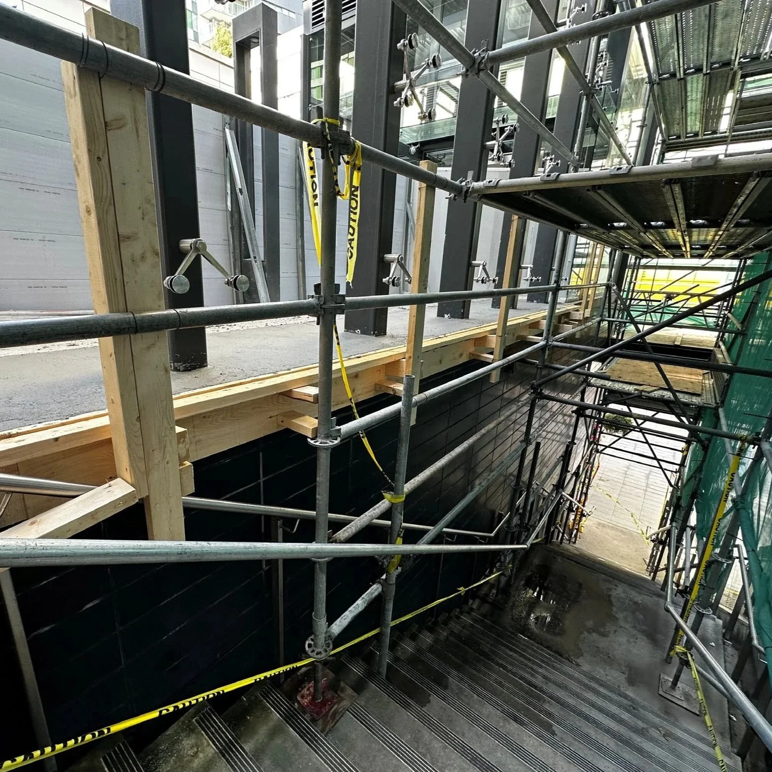 Construction site showing scaffolding and wooden framework alongside a building. A caution tape is visible, and the area appears to be under renovation or construction.