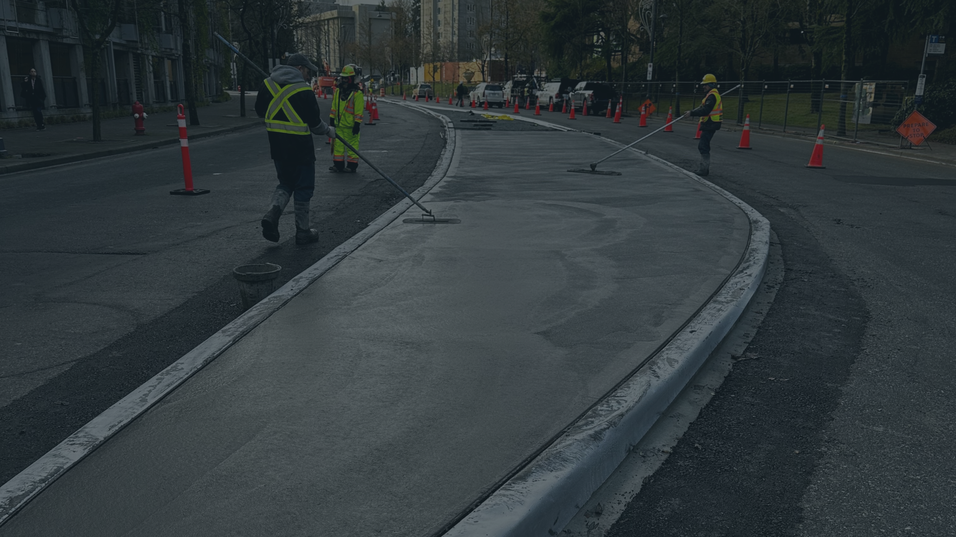 Construction workers in safety gear paving and smoothing a section of new concrete on a city street, with orange traffic cones and caution signs in the background.