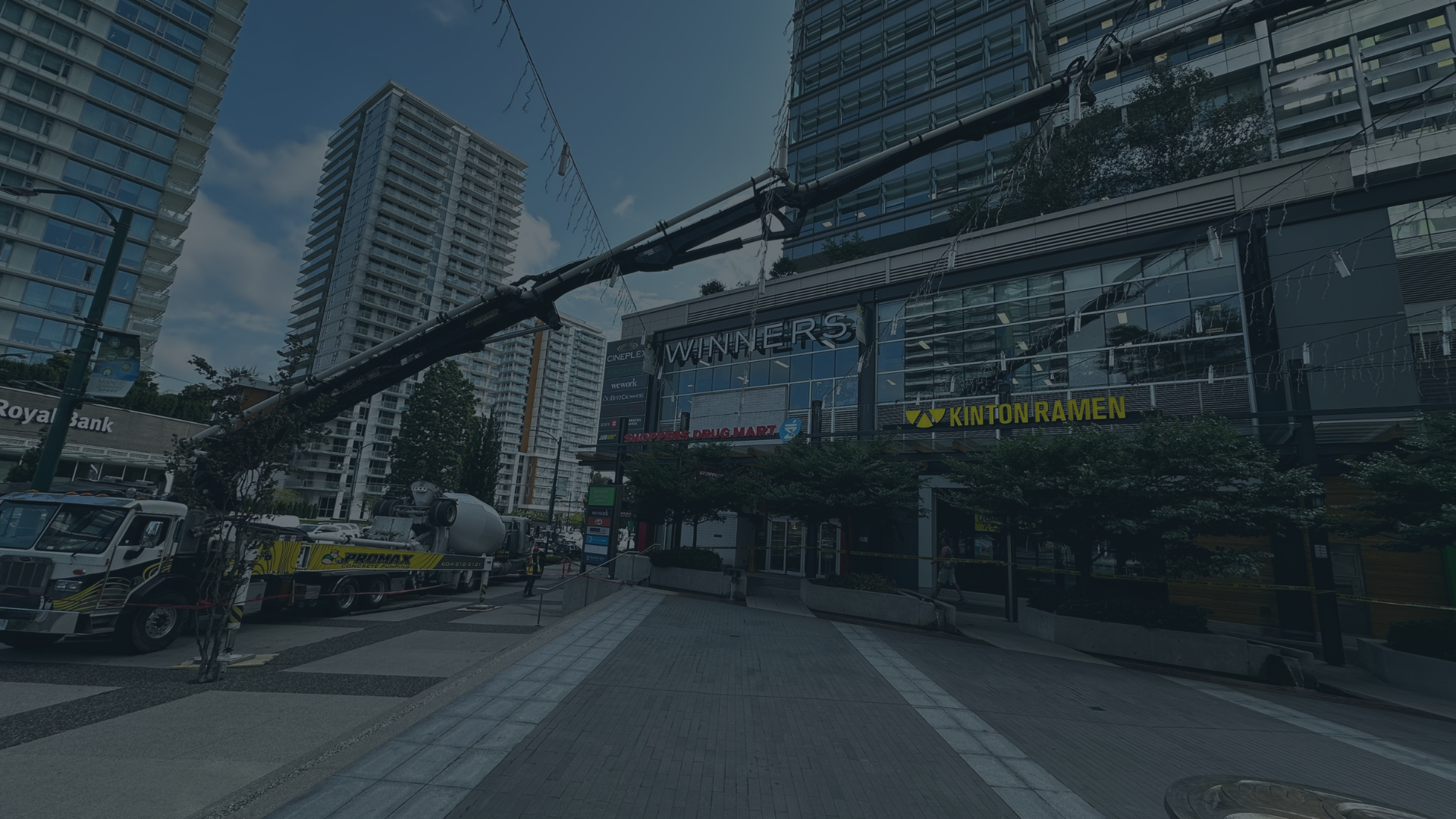 City street scene with tall modern buildings, a construction crane, and a truck parked on the side. There are signs for Winners, Shoppers Drug Mart, and Kinton Ramen. The sidewalk has trees and some pedestrians.