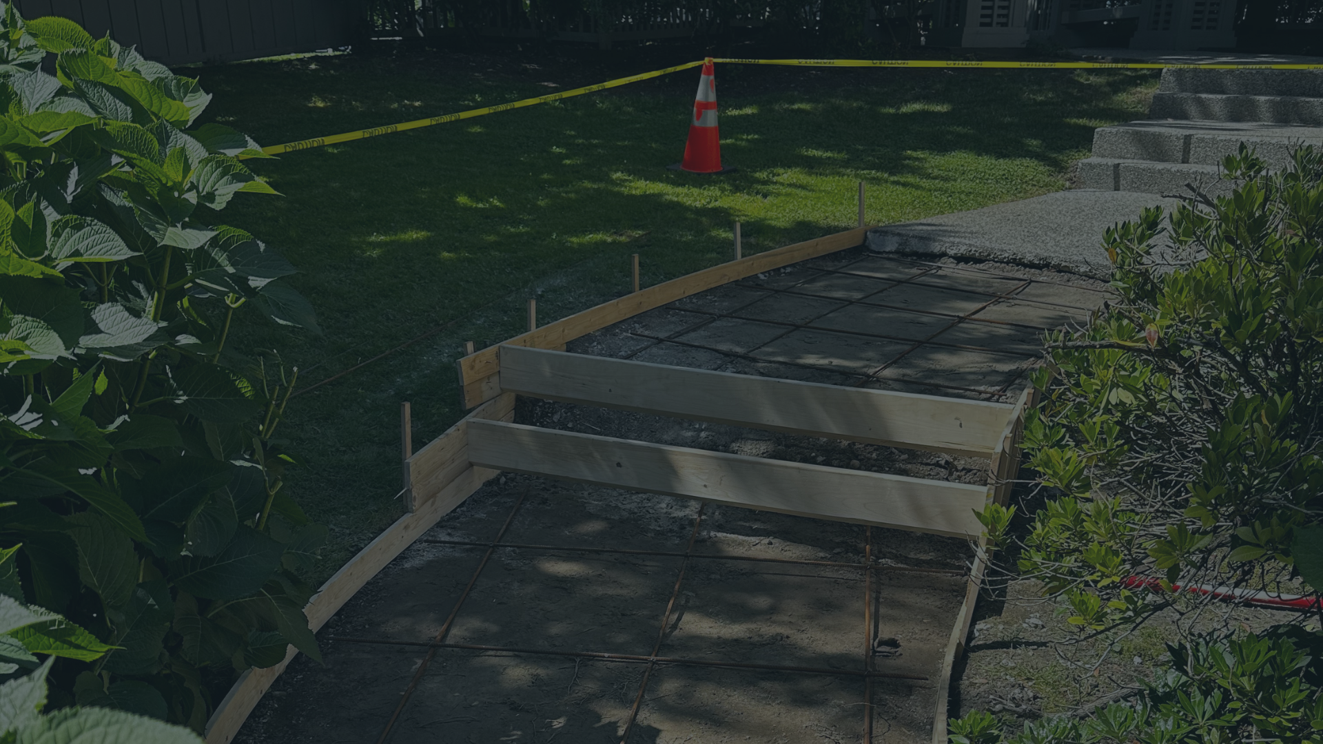 Construction site with a partially built concrete pathway, wooden framing, a safety cone, and caution tape, surrounded by plants and grass.