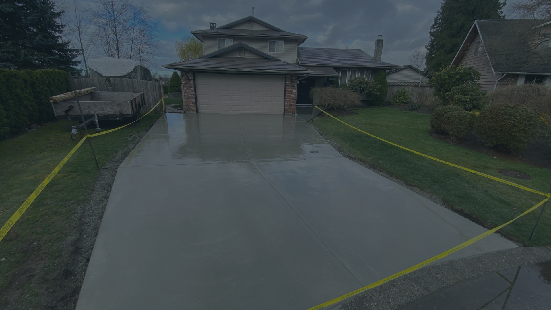 Wet driveway and lawn area with yellow caution tape surrounding the walkway, in front of a suburban house with a two-car garage, trees, the sky is cloudy.