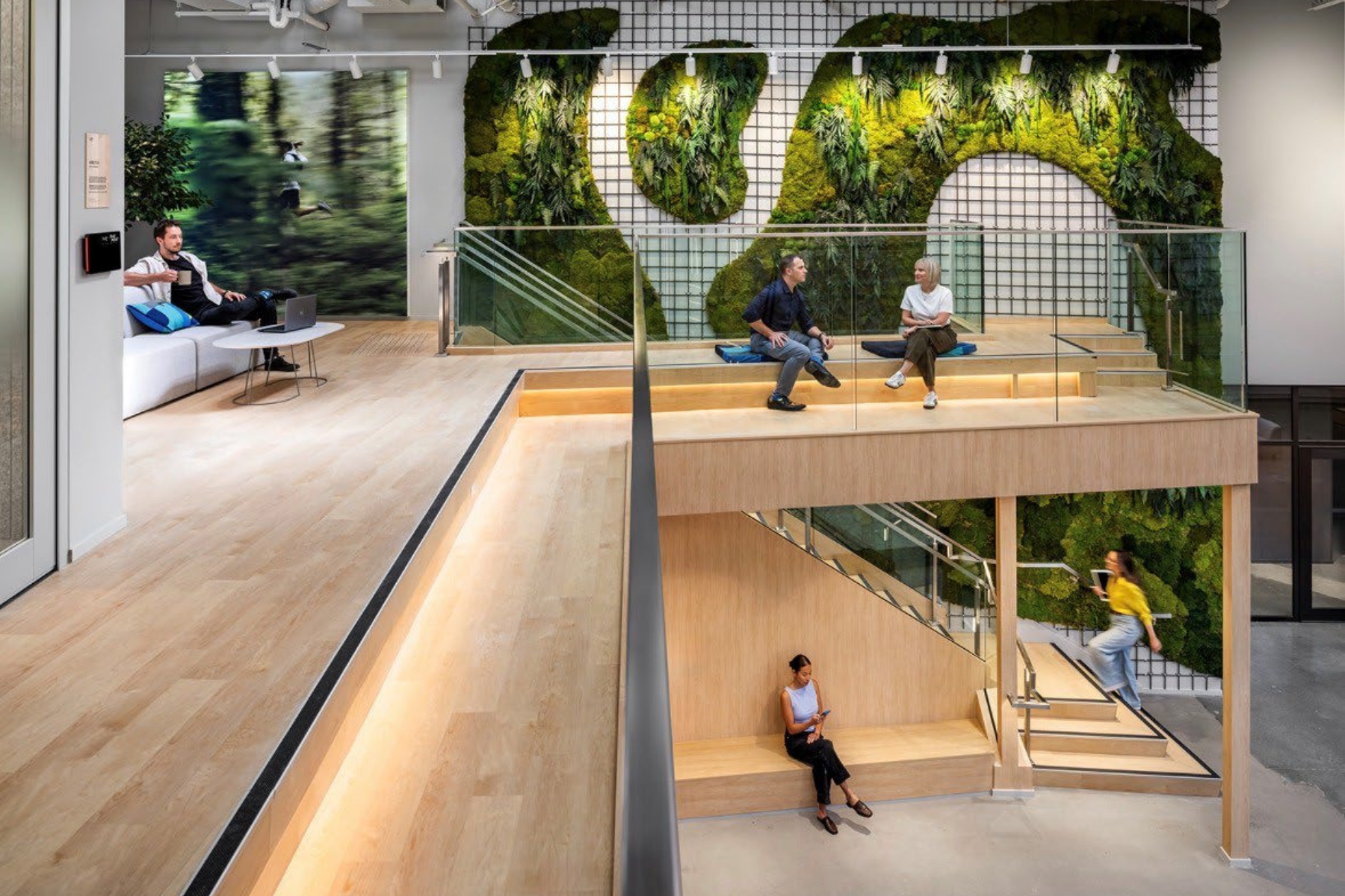 Modern office lounge with a green wall, people sitting on cushions and stairs, a woman using her phone, and a woman sitting on a bench, with wooden floors and glass railings.