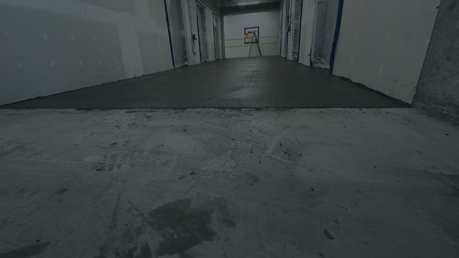 Interior view of a building under construction with a concrete floor, partially installed drywall, and a basketball hoop in the background.