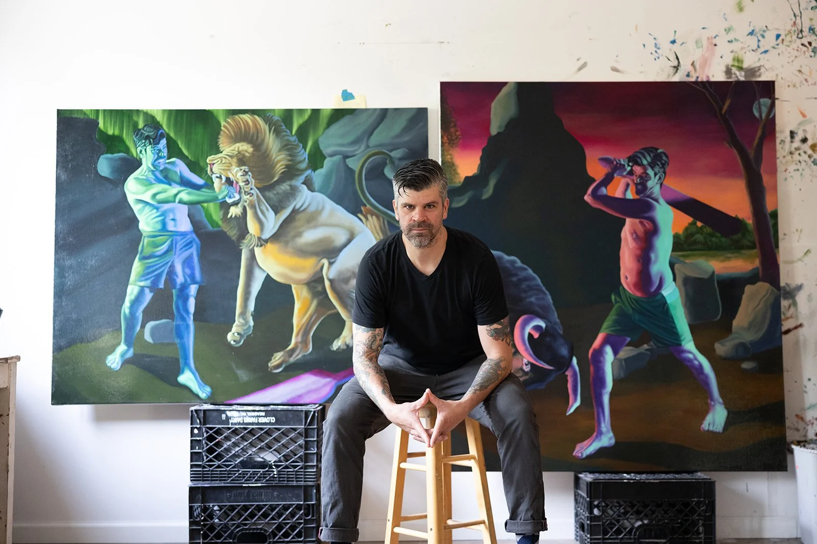 An artist with tattoos on his arms sitting on a stool in front of two large colorful fantasy landscape paintings, one featuring mythical creatures and the other a sunset scene with a person and a tree.