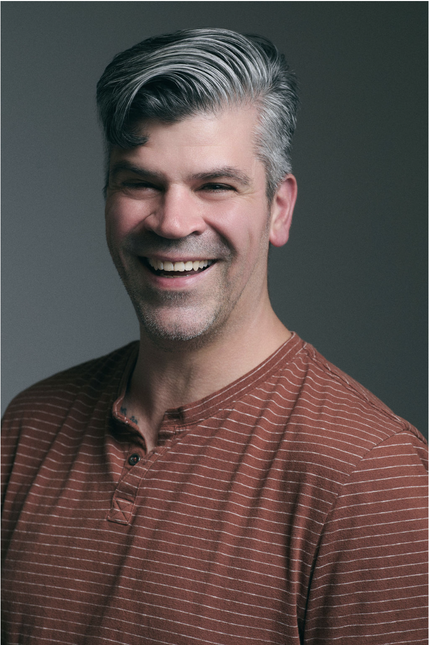 A smiling middle-aged man with gray hair and stubble, wearing a rust-colored, striped, buttoned shirt against a dark gray background.
