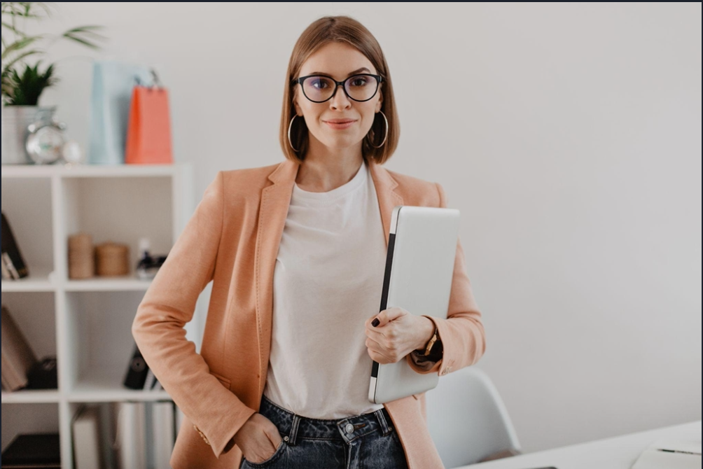 A woman with glasses and hoop earrings holding a closed laptop while standing in an office.