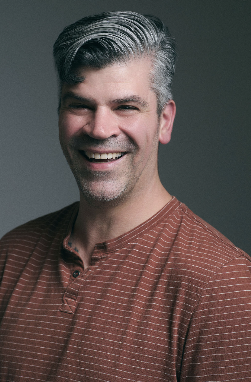 A smiling middle-aged man with gray hair, wearing a rust-colored, striped shirt against a gray background.