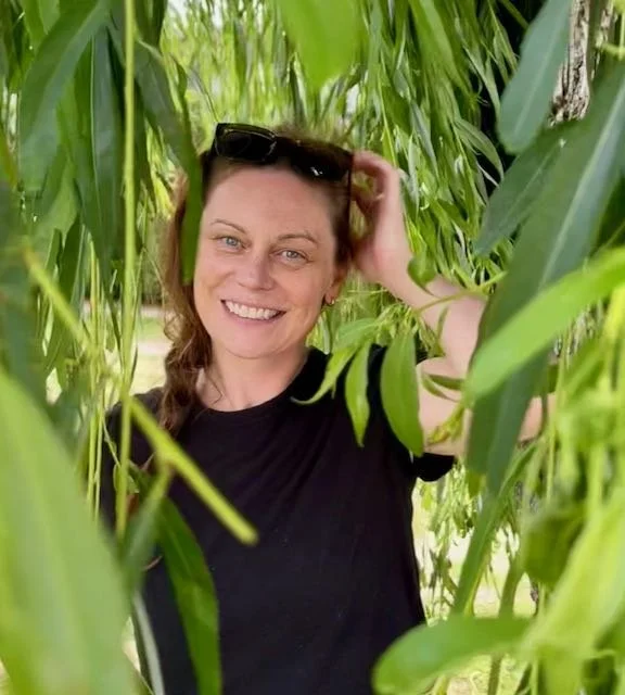 Woman smiling and standing among tall green plants or corn stalks outdoors.