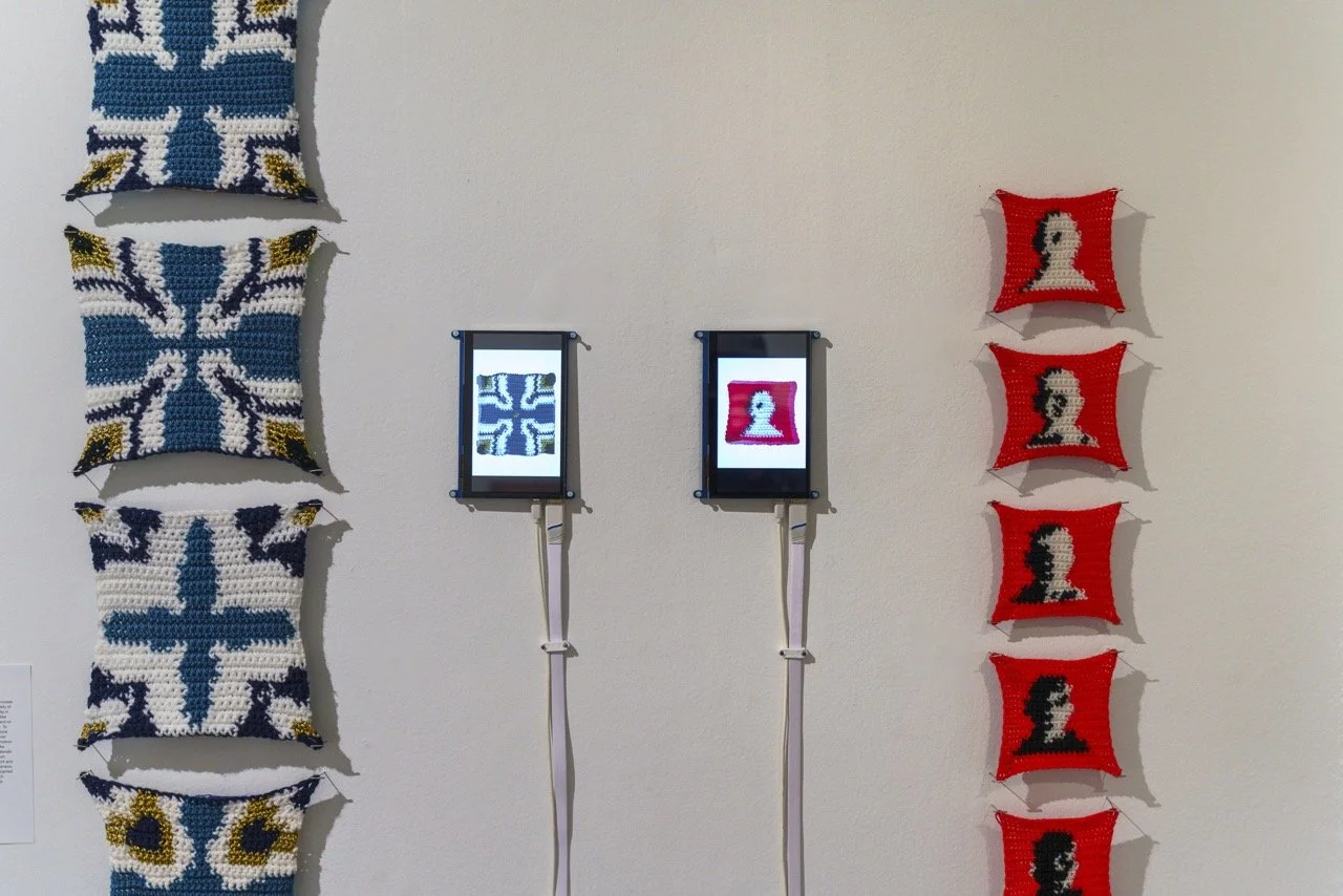 An art installation with knitted pillows on a white wall, two digital screens in frames displaying pixel art images, and a row of pixel art pillows on the right side.