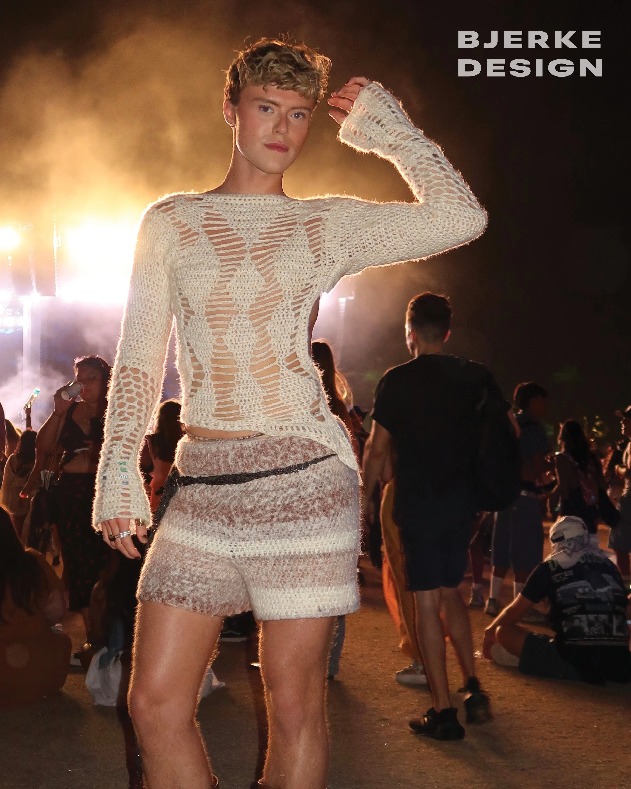 A woman in a beige crochet dress with shorts stands in a crowd at night, backlit by bright lights, with other people around her.