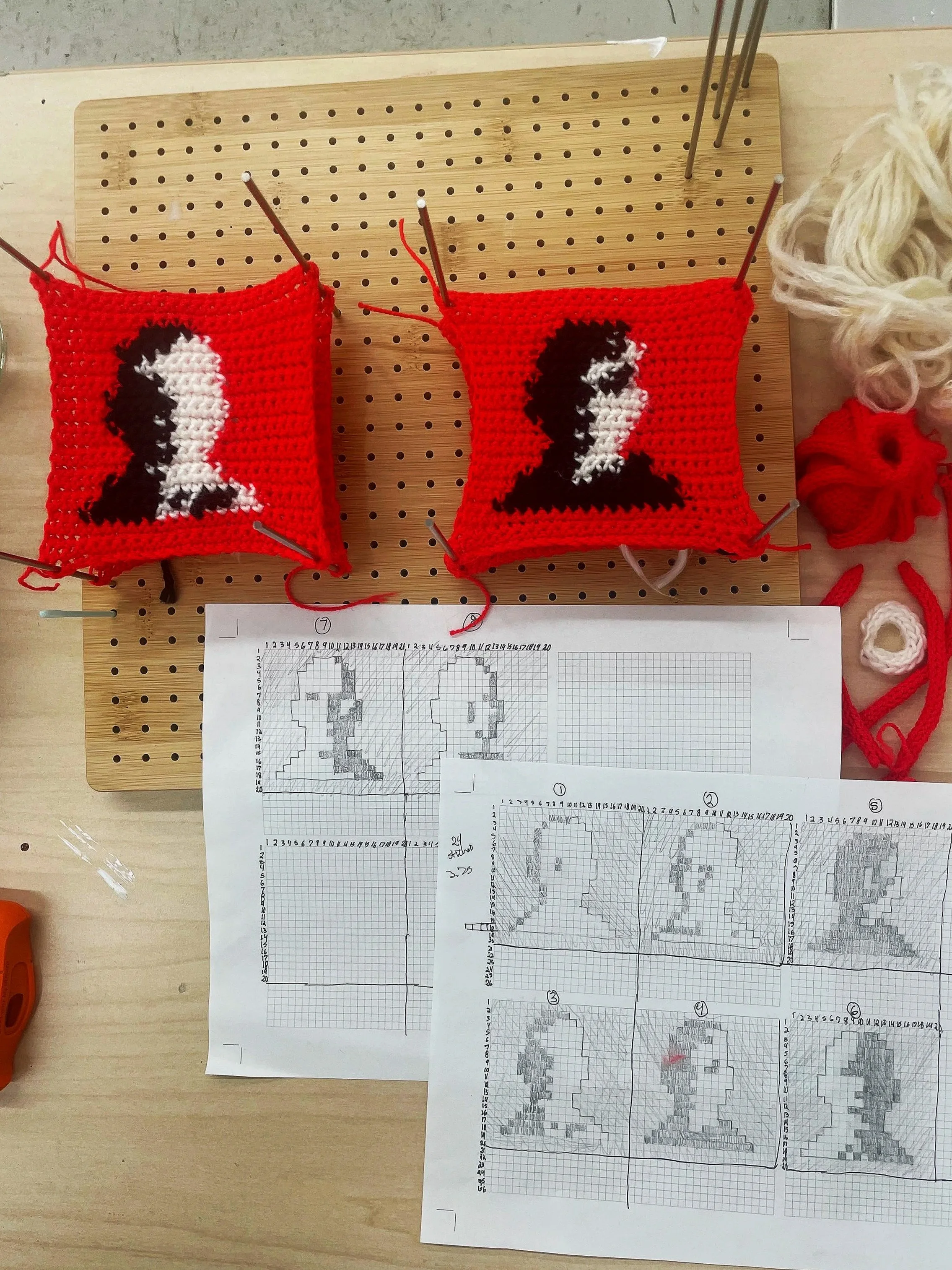 Two crocheted pieces with a portrait of a man with dark hair, placed on a wooden crafting board. The pieces are red with a black and white face in the center. To the right, there are yarn balls and crochet tools, and below the pieces, there are printed blueprint patterns for the design.