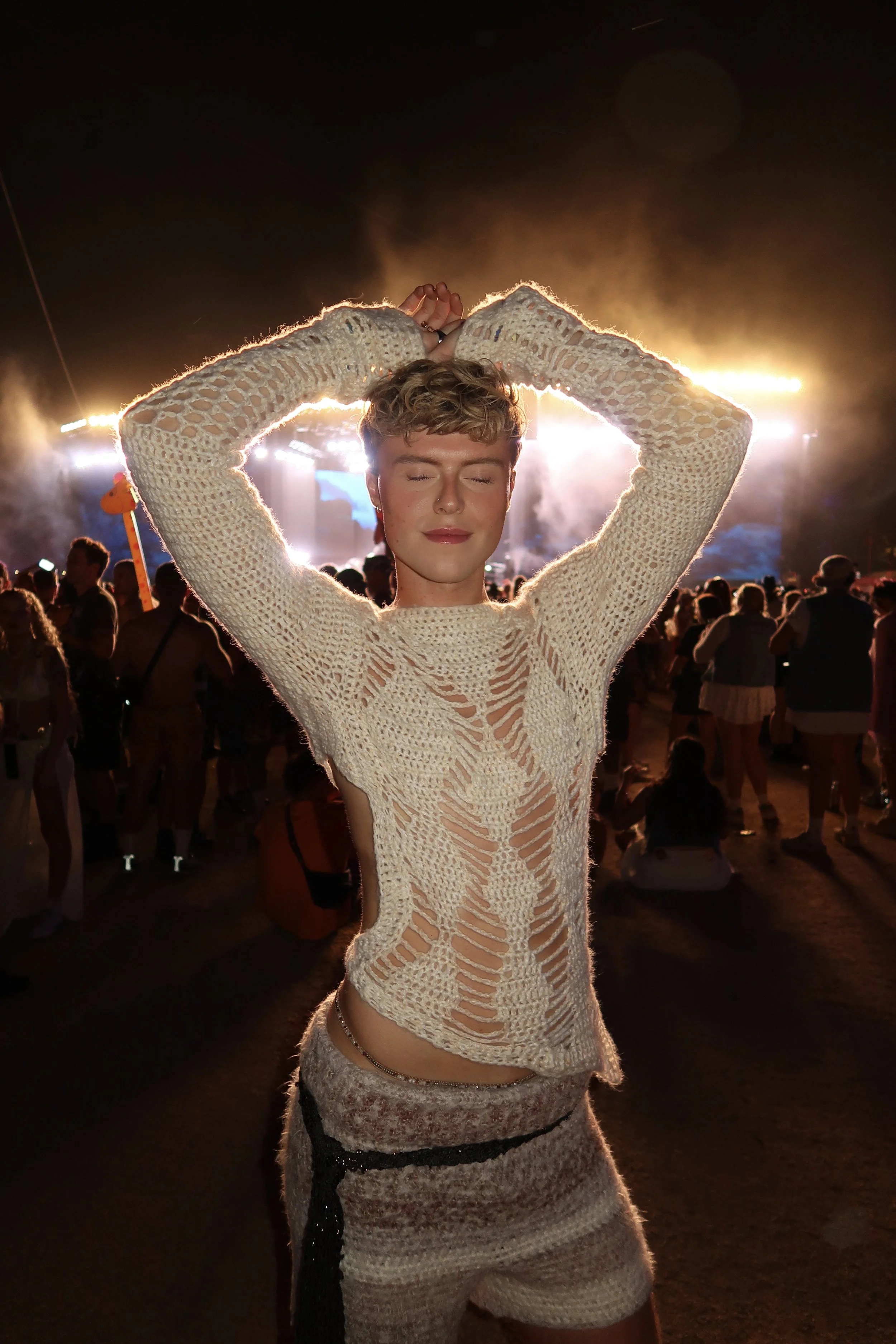A woman with short, curly blonde hair and closed eyes is standing with her arms raised above her head in a heart shape at a nighttime outdoor music festival. She is wearing a cream-colored, open-knit crochet top and gray shorts. The stage with bright lights and a crowd of people are visible in the background.