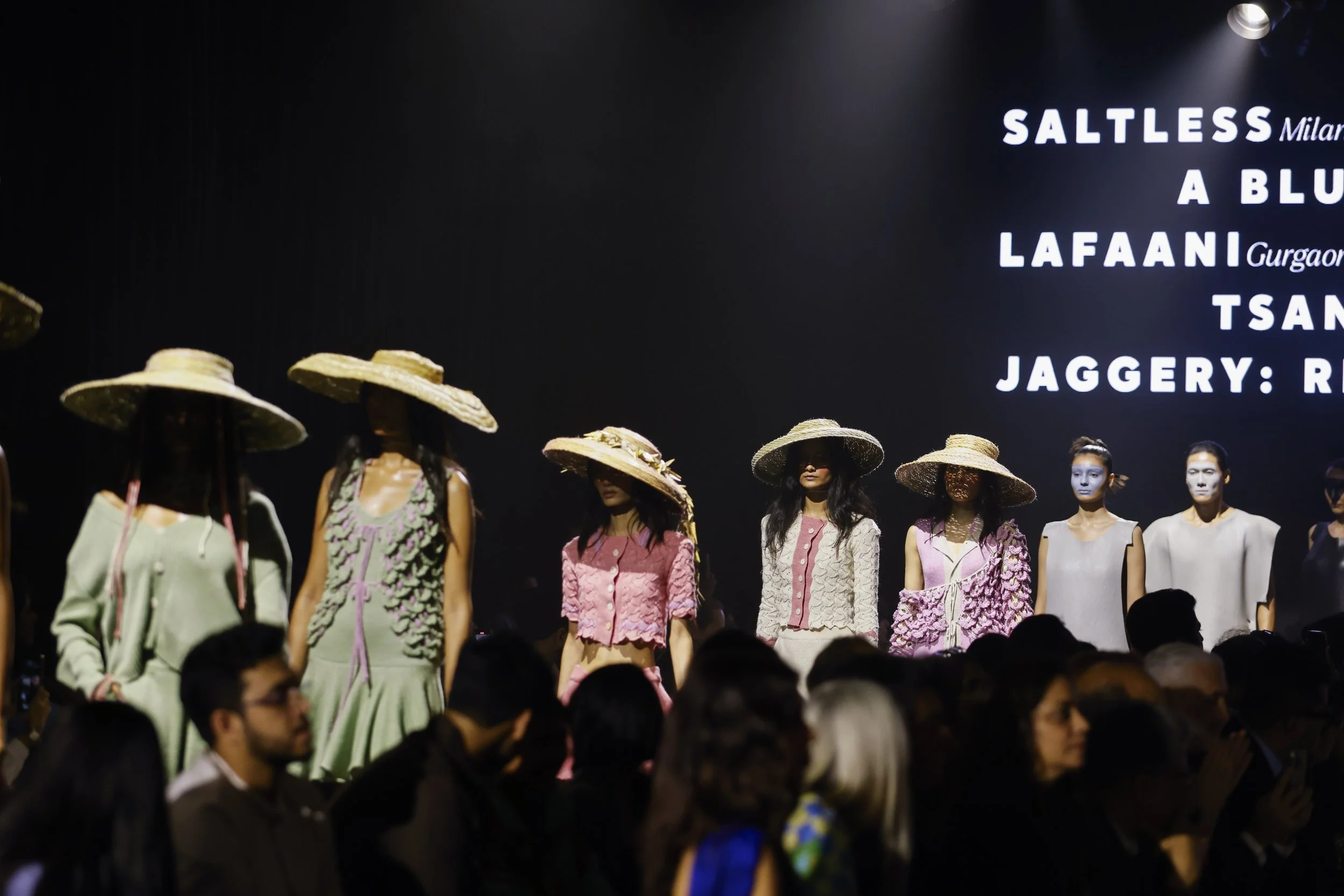 Fashion models on runway wearing pastel-colored outfits and large sun hats, with audience in foreground and text on a screen behind them.
