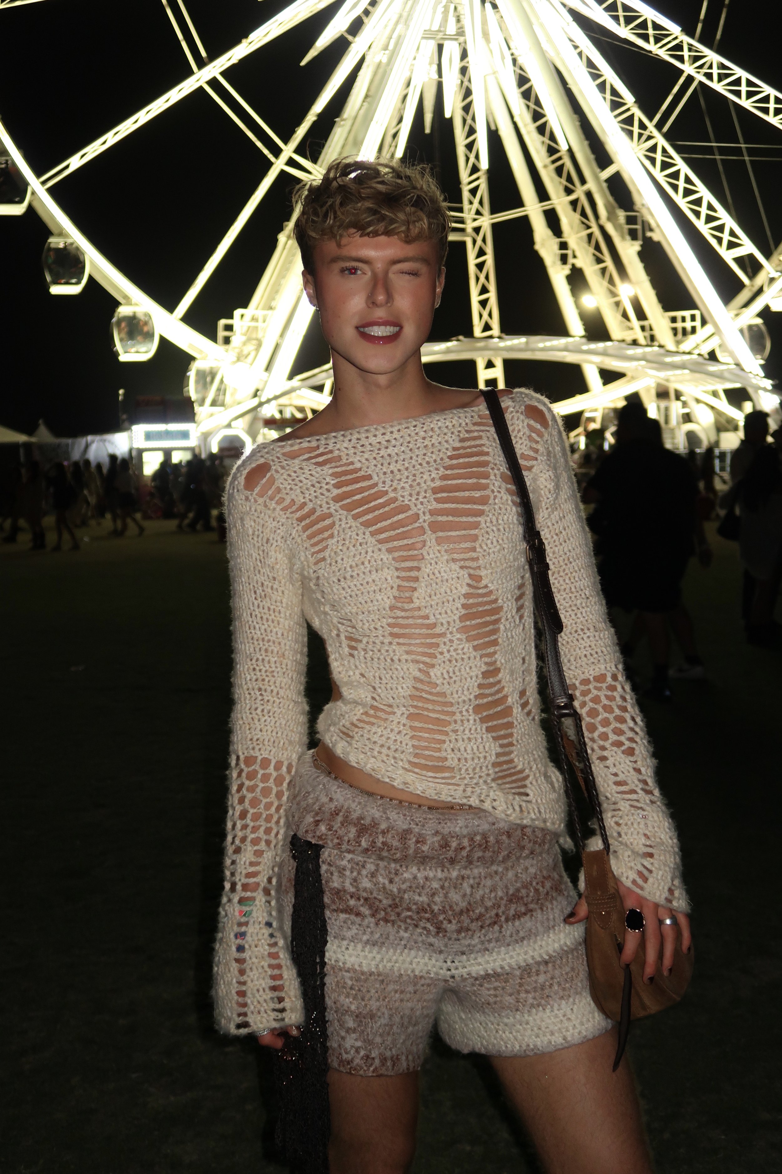 A woman with short curly hair and a sleeveless beige crochet top at a night carnival with a brightly lit Ferris wheel in the background. She is winking and smiling slightly, holding a brown purse with a shoulder strap.