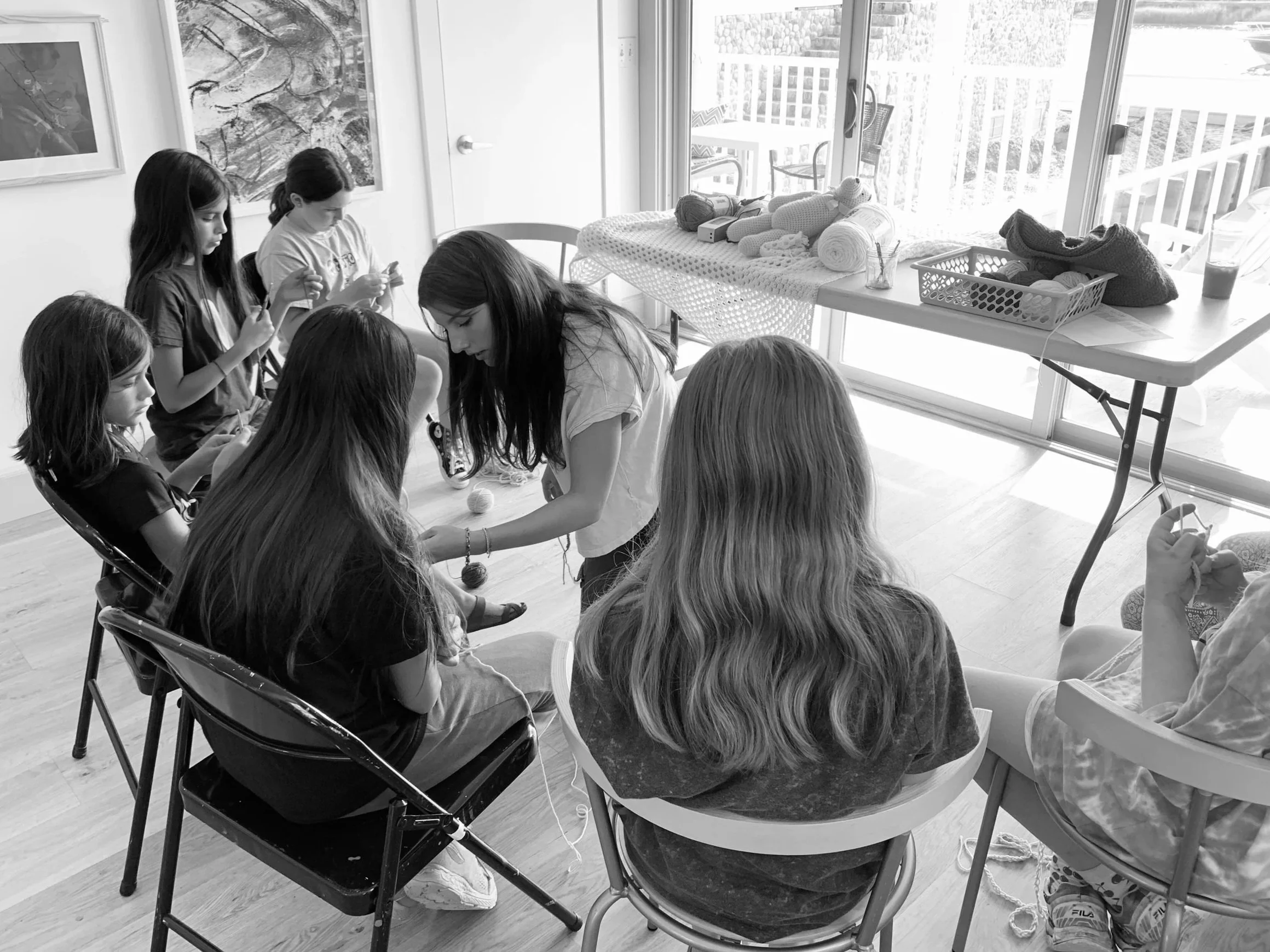 A group of young girls sitting on chairs in a room, working on a knitting or craft project, with a table nearby holding yarn, knitting supplies, and an art piece on the wall, with a sliding glass door leading to an outdoor balcony.