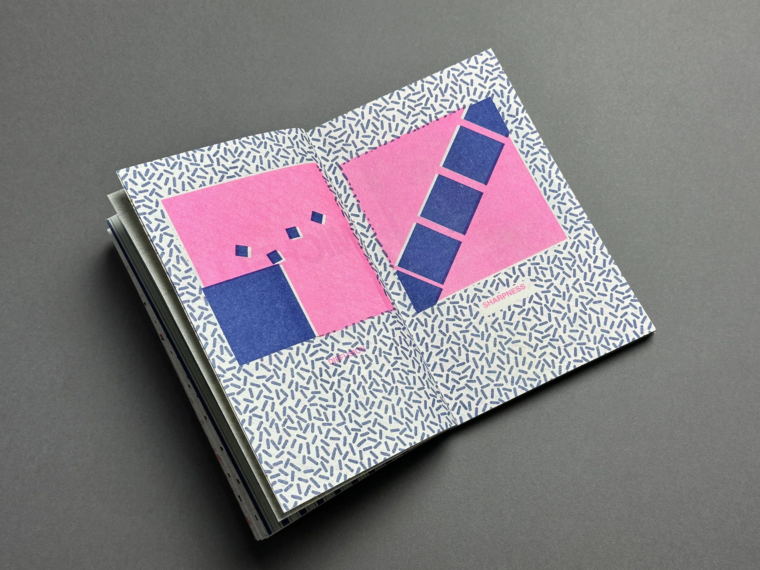 An open book with pages containing graphic design elements, including pink backgrounds, blue rectangles, and a pattern of small curved lines, on a gray surface.