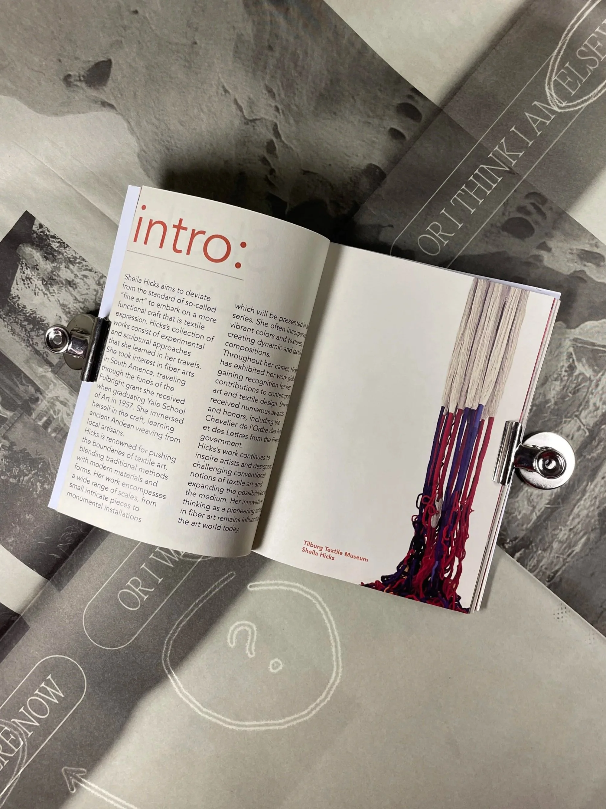 Open booklet with an article about Sheila Hicks, a textile artist, featuring a color photograph of woven textile art hanging from the right page, beneath a silver clip. The background surface is a smooth, gray material with words and arrow patterns.