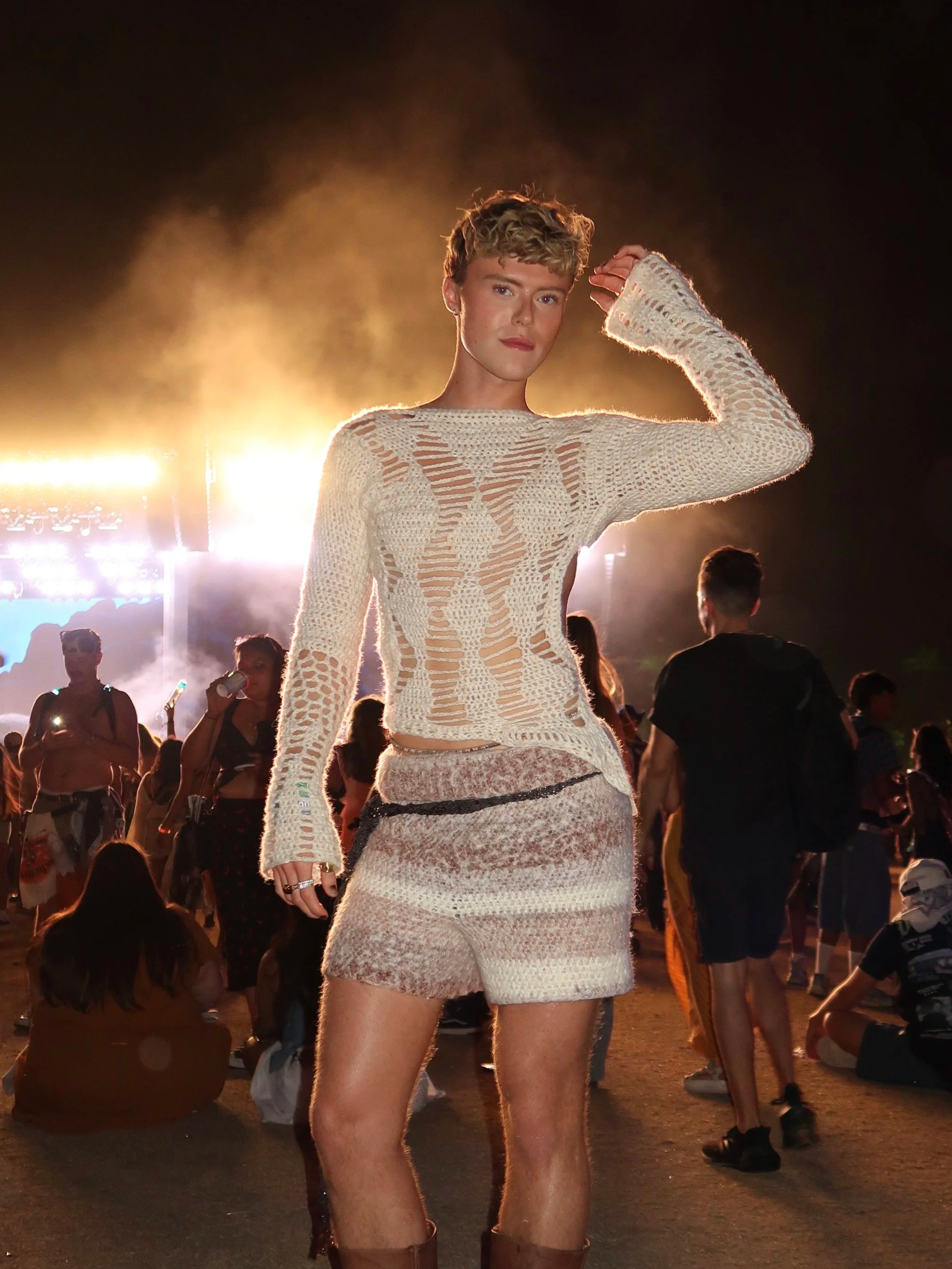 A woman standing in a crowd at night, wearing a beige, distressed knit outfit with shorts and tall boots, with stage lights behind her.