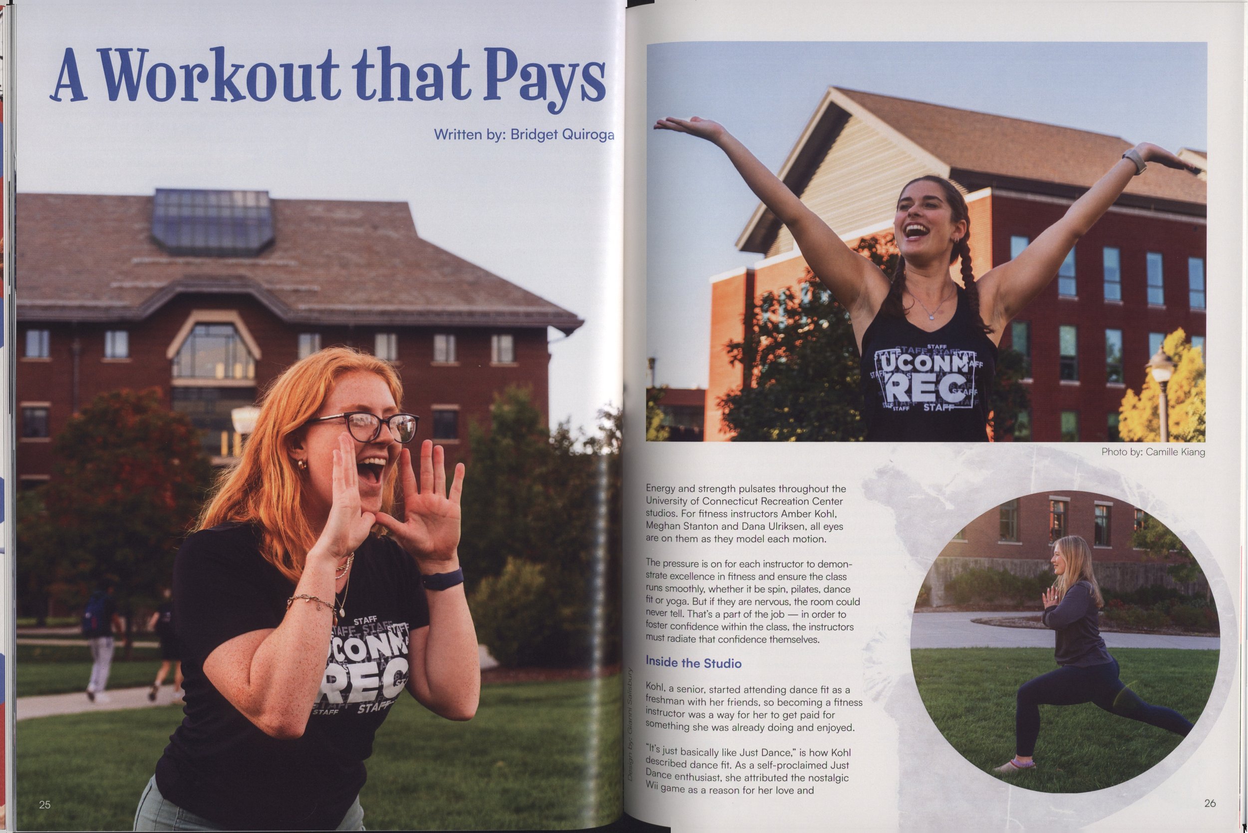 A woman with long red hair and glasses, wearing a black t-shirt with the words 'UCONN REG' and 'STAFF' on it, is smiling and making a heart shape with her hands. In the background, there's a woman with braided hair and a black tank top, with her arms raised, and a large brick university building with a brown roof and multiple windows. The scene takes place outdoors on a grassy area with trees and a walkway.