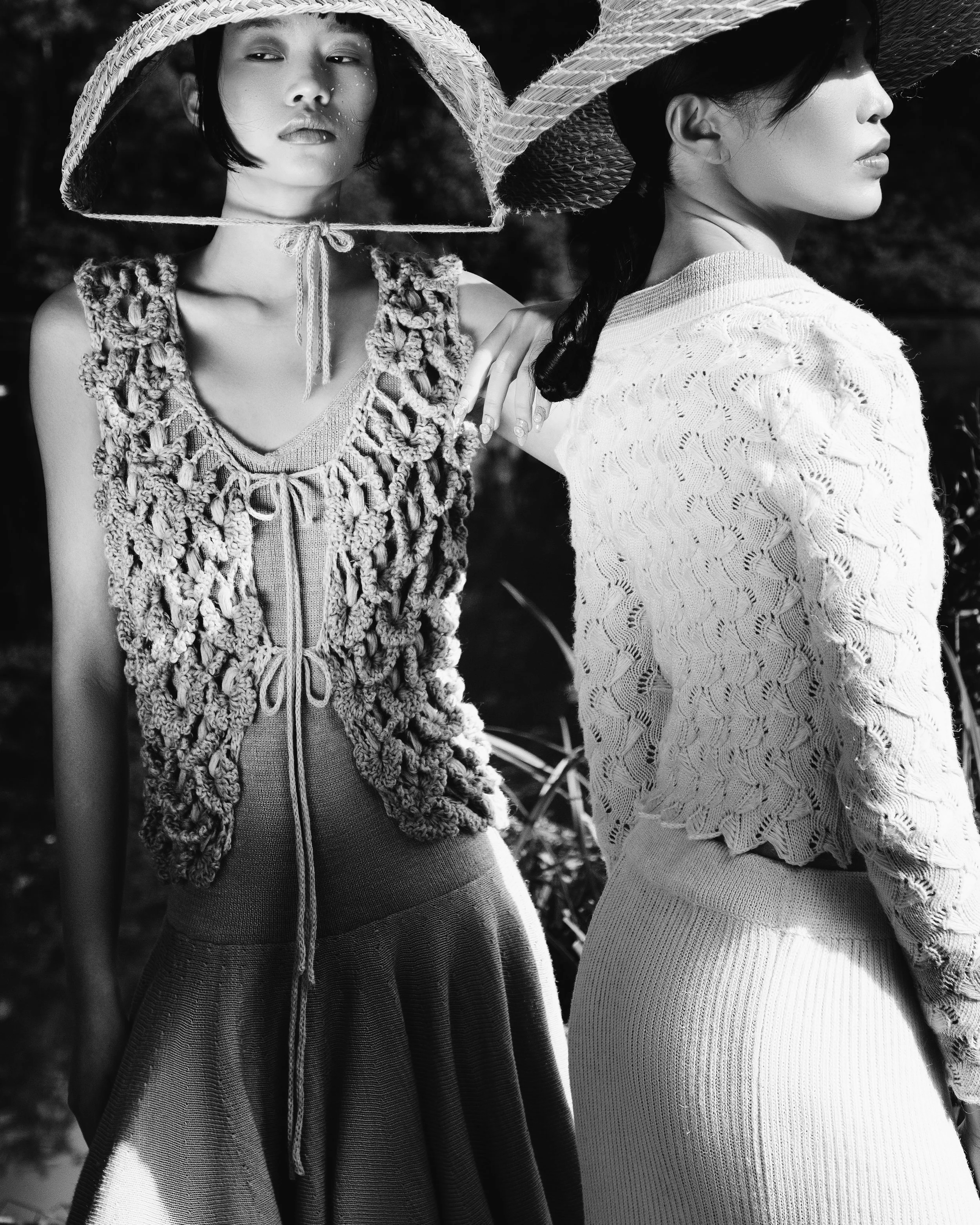 Two women wearing wide-brimmed straw hats, standing close together outdoors, with one woman's hand resting on the other's shoulder. The photo is in black and white, focusing on their profiles and clothing textures.