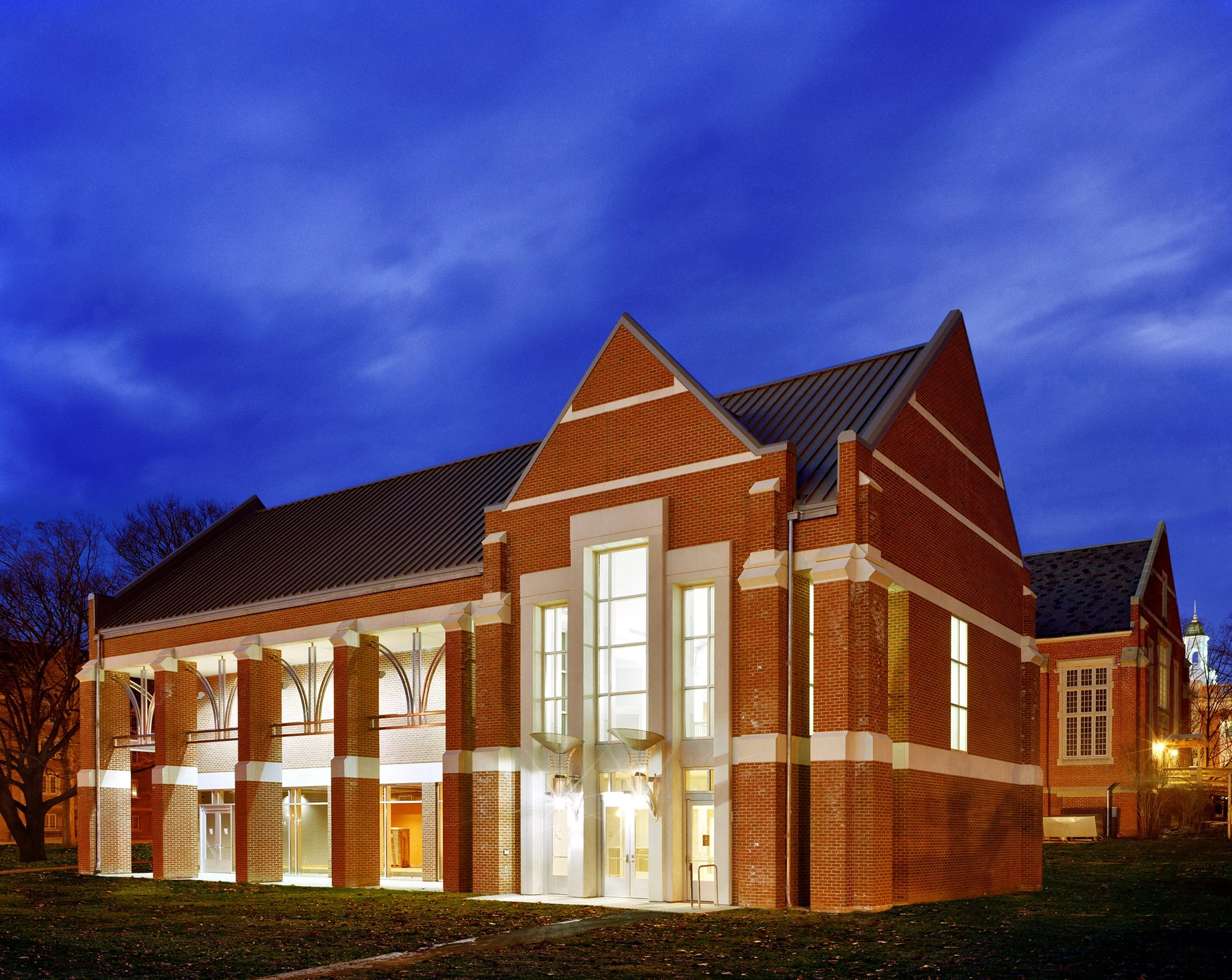 A brick building with large illuminated windows and a peaked roof at dusk, with decorative arches supported by columns at the entrance and trees in the background.