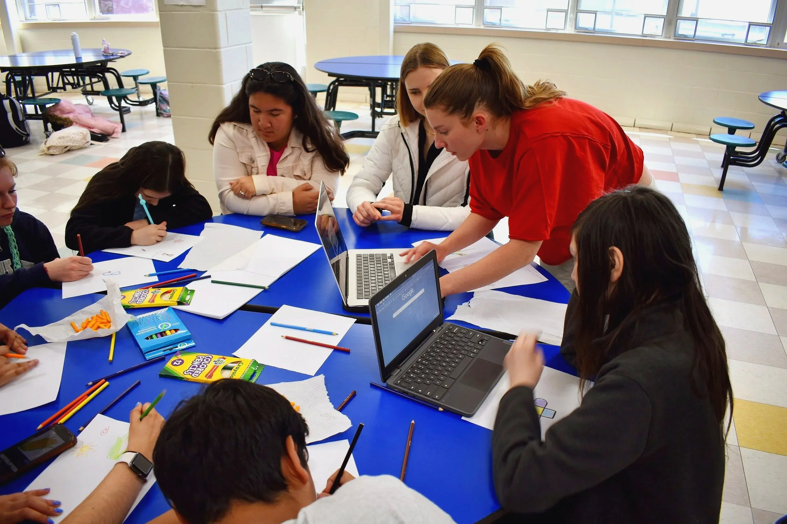 Group of children and a teacher gathered around a blue table in a school cafeteria, engaged in a learning activity with paper, colored pencils, and laptops.