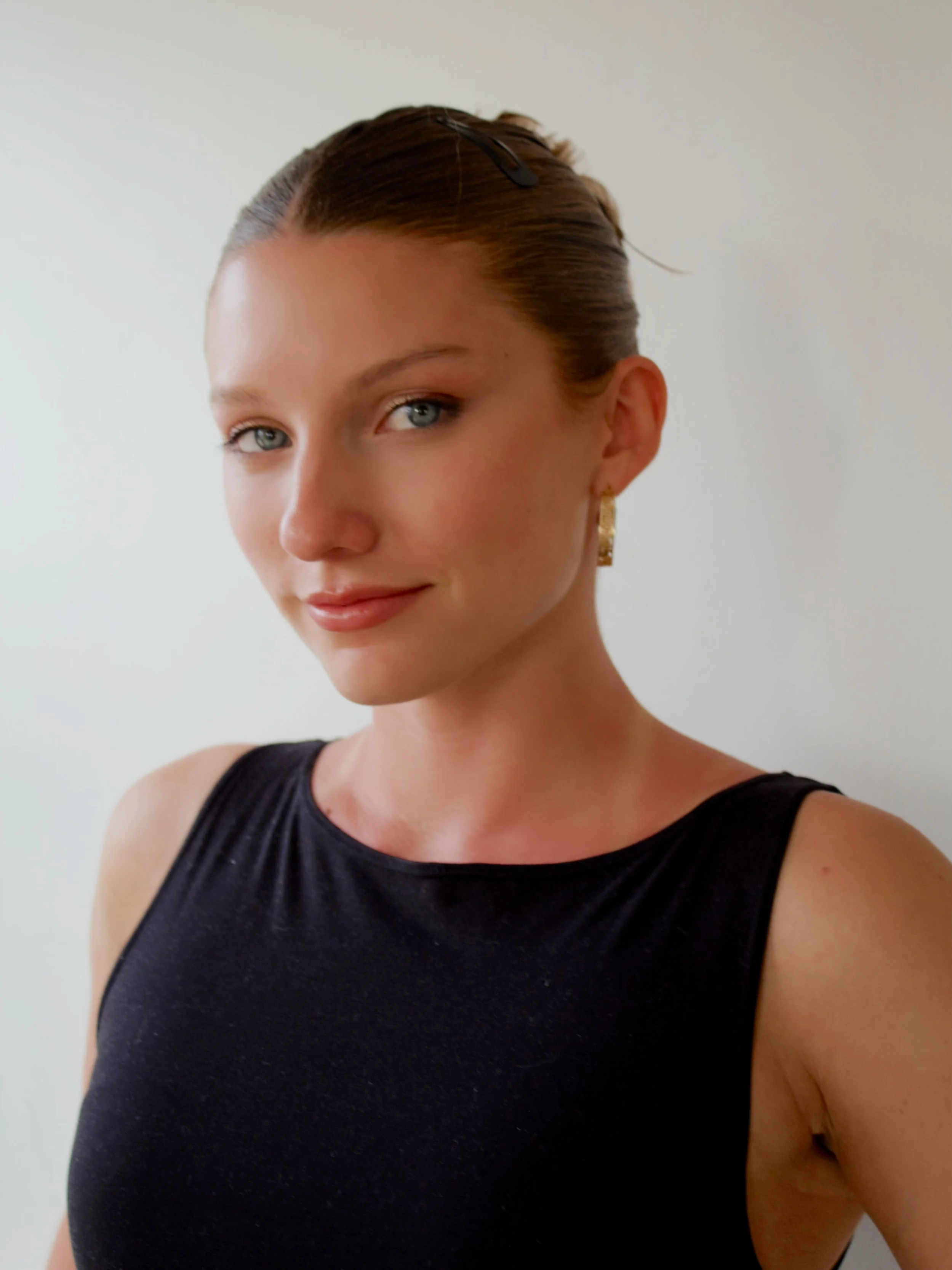 A woman with fair skin and blue eyes wearing a black sleeveless top, gold earrings, and a hair clip, smiling slightly, standing against a plain light-colored wall.