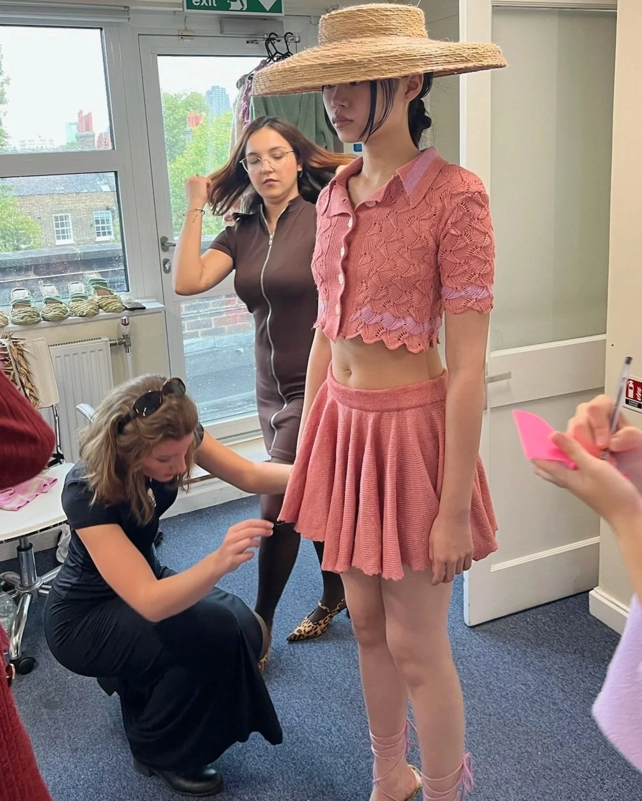 A woman in a pink lace crop top and matching skirt is standing in a room with her eyes closed, wearing a large woven hat that covers her eyes. Another woman is kneeling on the floor adjusting her outfit, and a woman with glasses and brown hair is standing nearby, adjusting her hair.