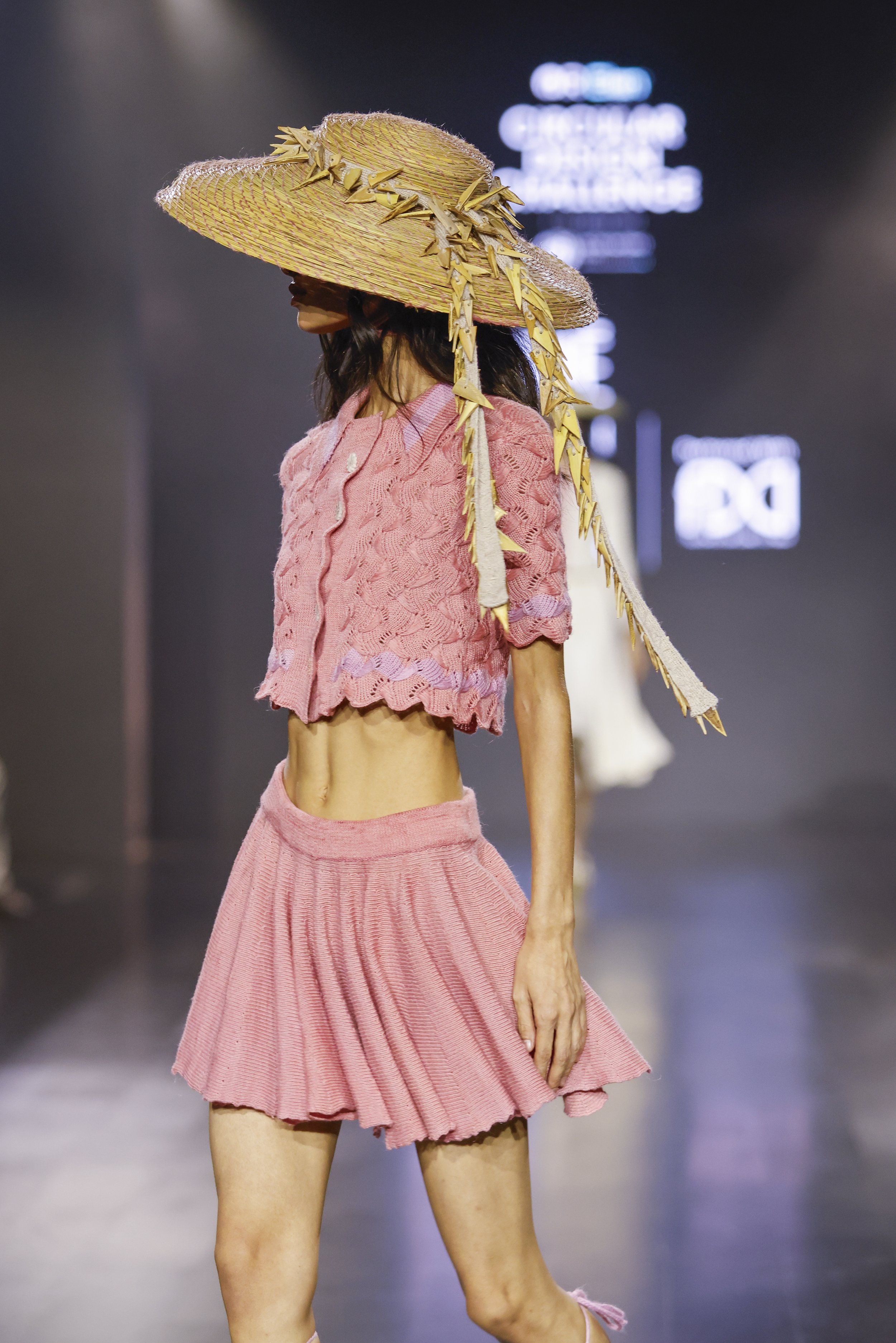Fashion model walking on runway wearing pink knit top with scalloped edges, matching pink pleated skirt, and oversized straw hat with decorative yellow elements.