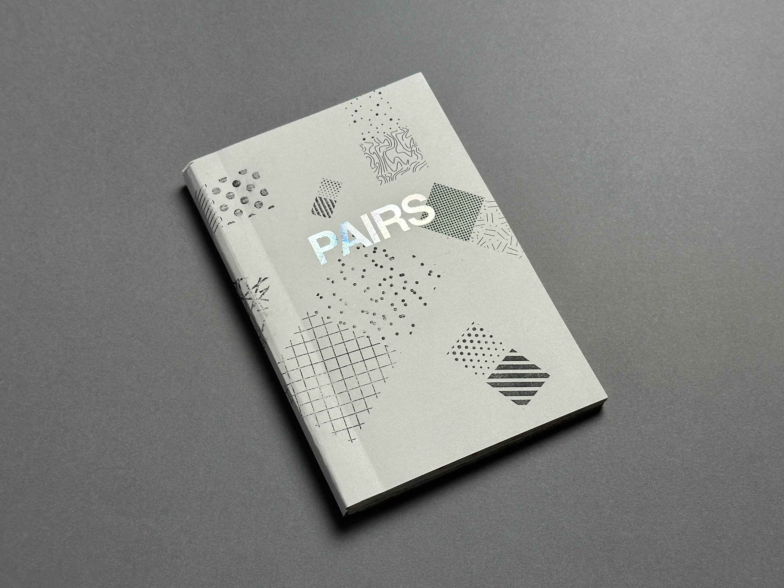 A white booklet with abstract black patterns and the word 'PAIRS' on the cover, resting on a gray surface.